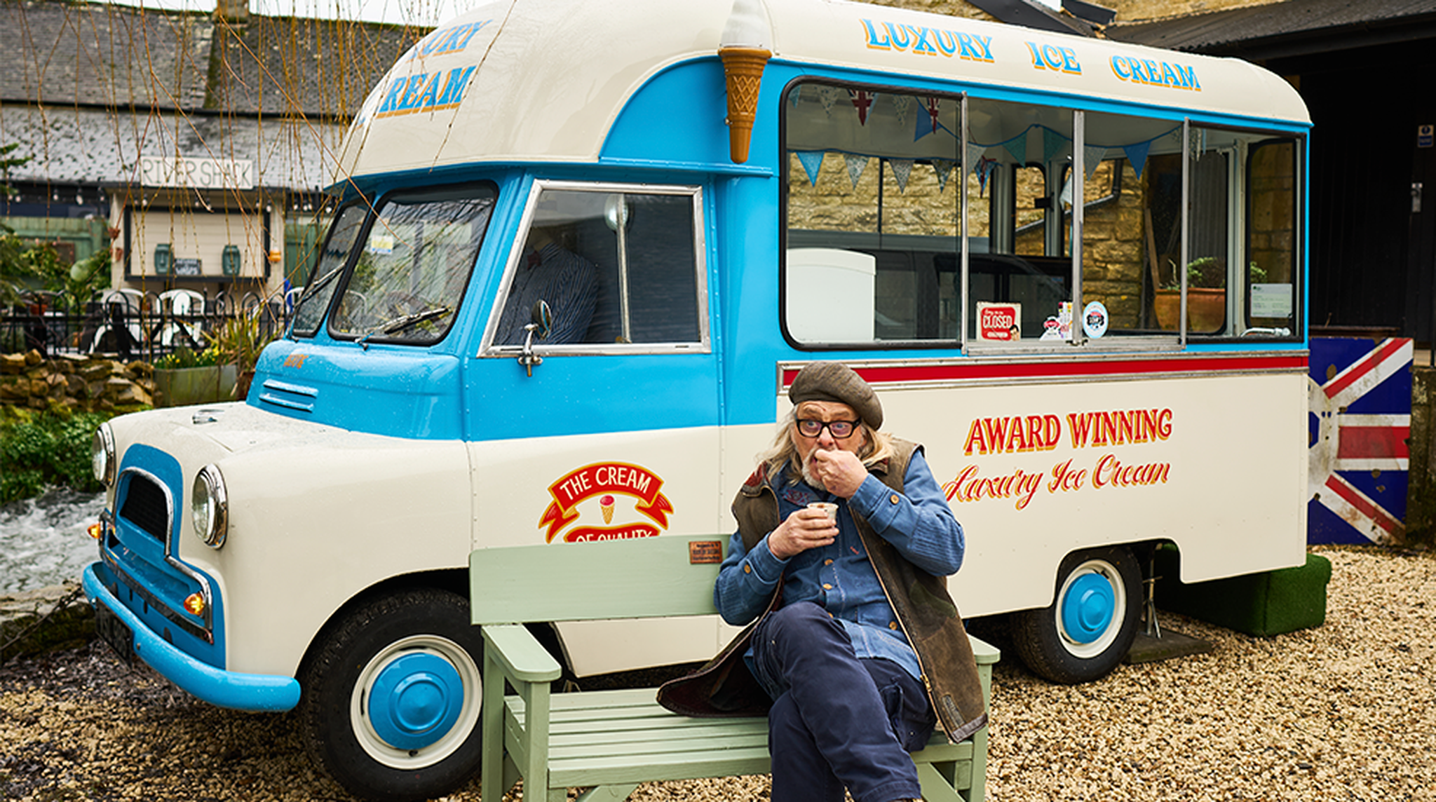 A person sits on a bench enjoying ice cream in front of a retro blue and white ice cream truck with "Award Winning Luxury Ice Cream" written on it.