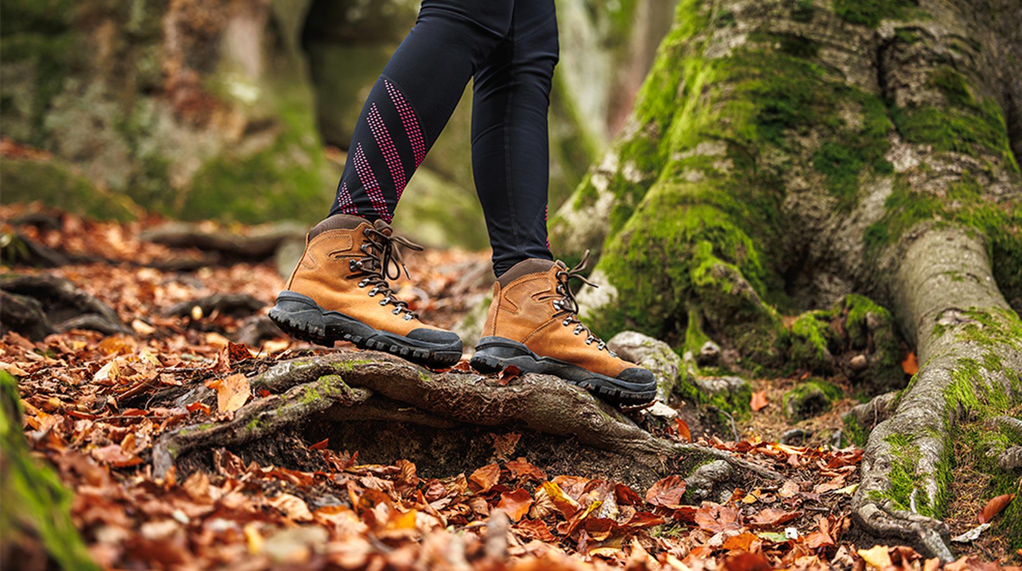 Person in brown hiking boots and black leggings standing on tree roots amidst fallen autumn leaves in a forest.