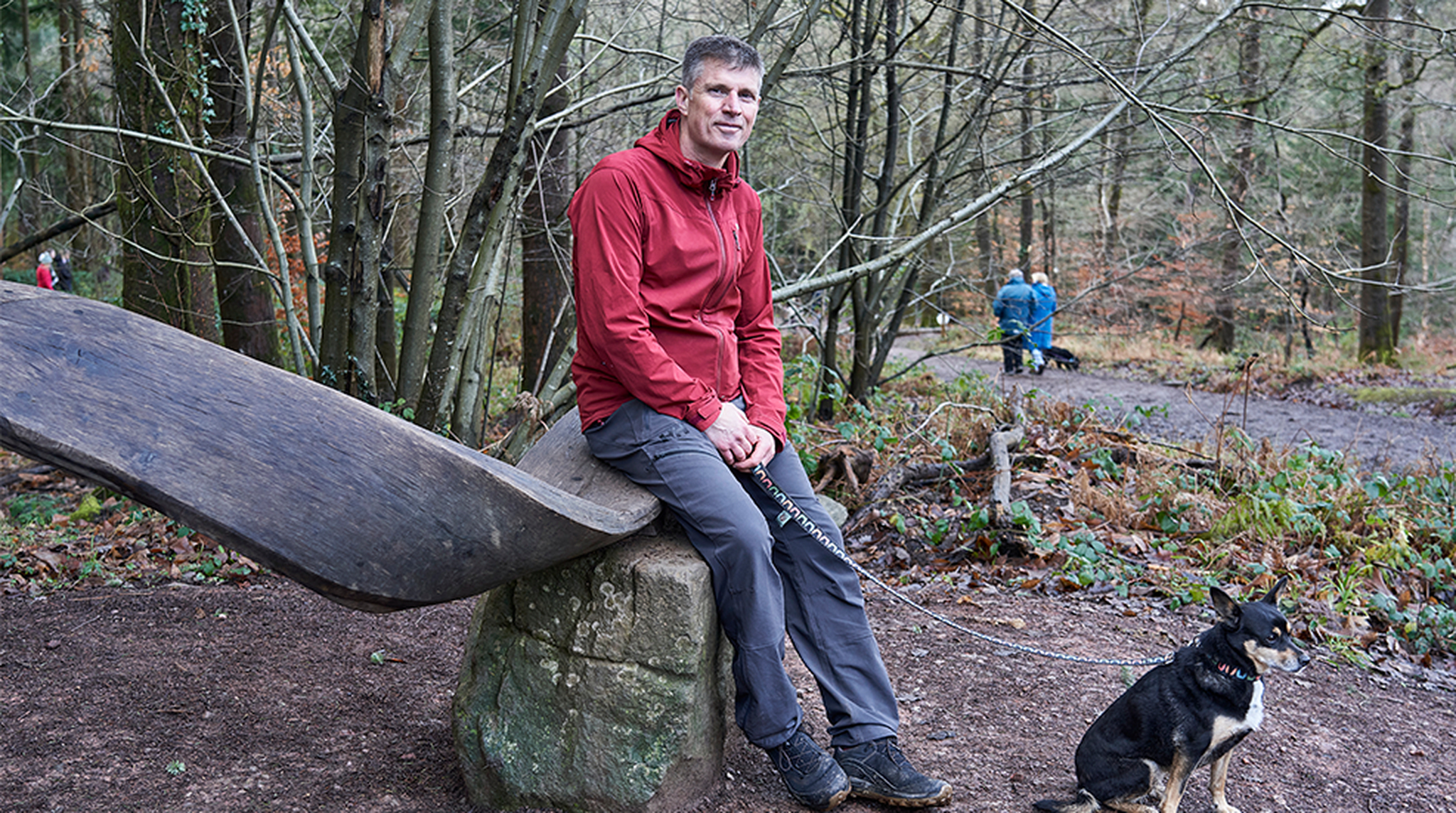 Man in a red jacket sitting on a wooden seat in a forest with a dog on a leash nearby, surrounded by trees and fallen leaves.