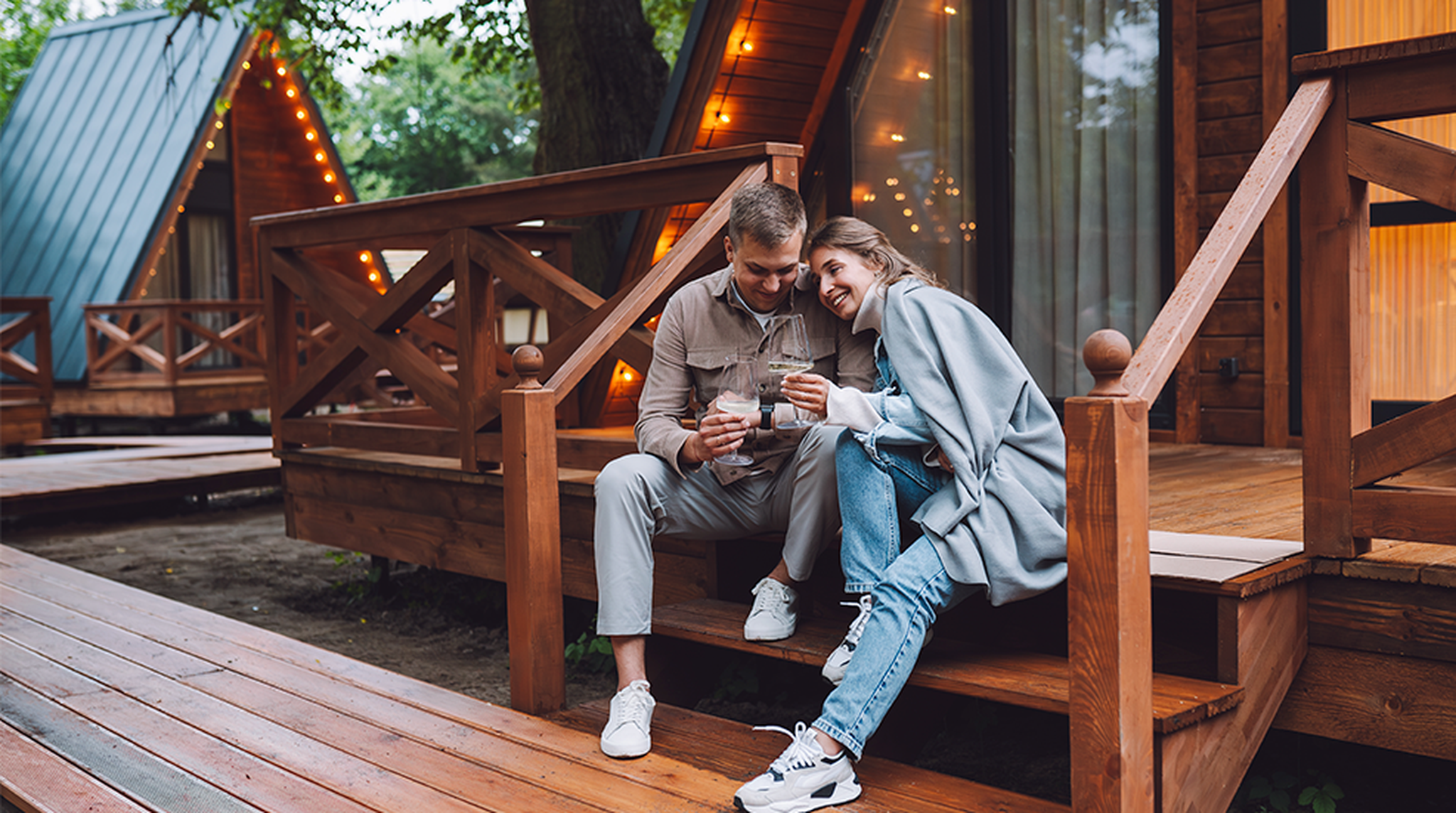 A couple sits on a wooden porch of cabin, smiling and holding drinks