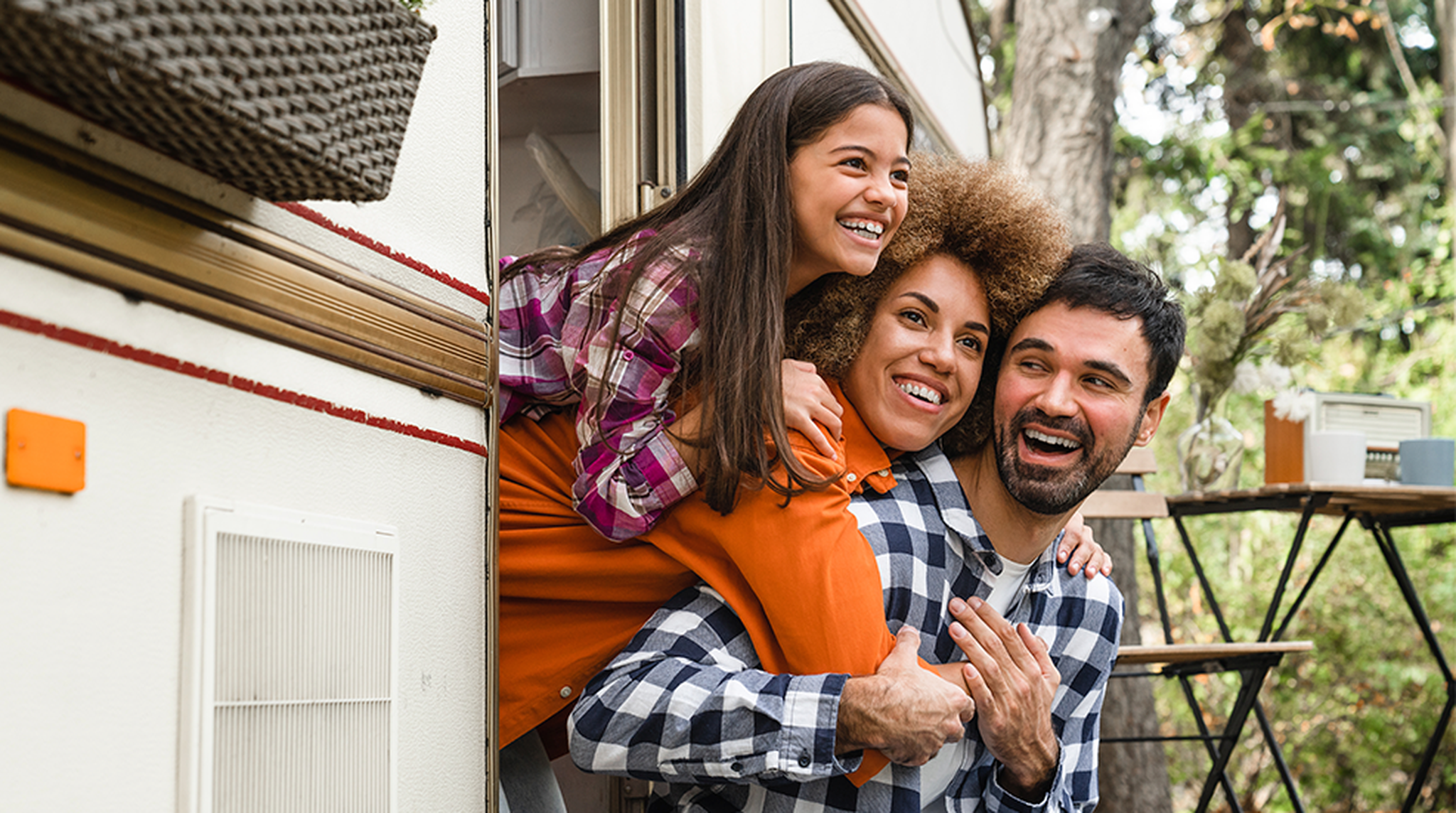 Family joyfully leans out of a camper door, all wearing plaid shirts. Trees in the background hint at a camping scene.