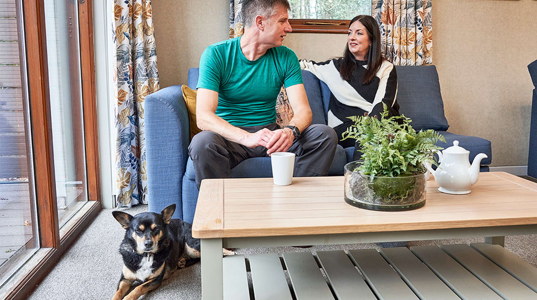 A couple sits on a couch by a window, talking and smiling. A black and tan dog lies on the floor. A table with a plant and teapot is in front.