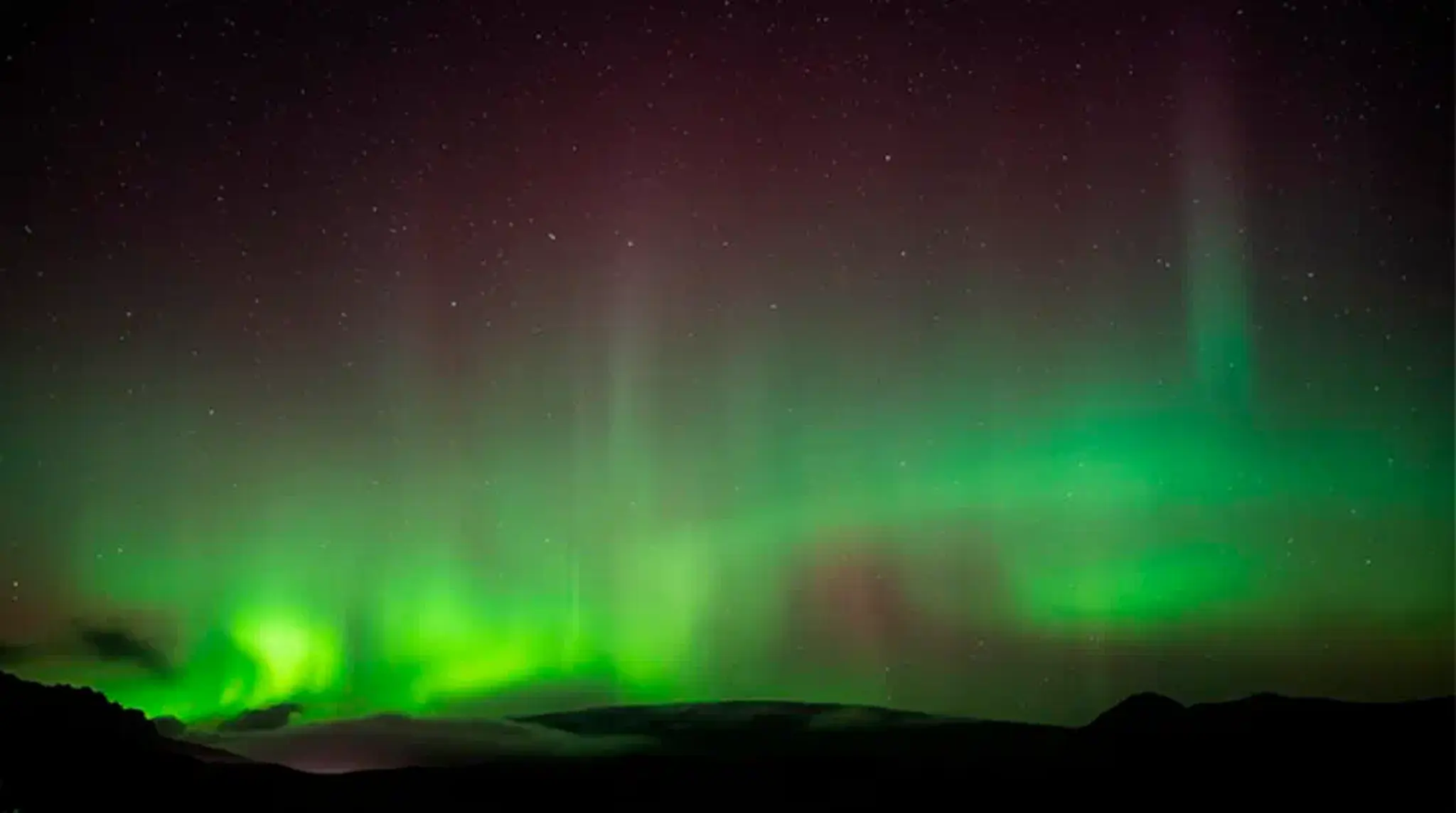 The image shows a vibrant aurora borealis with green and purple hues lighting up a star-filled night sky above dark silhouettes of hills.