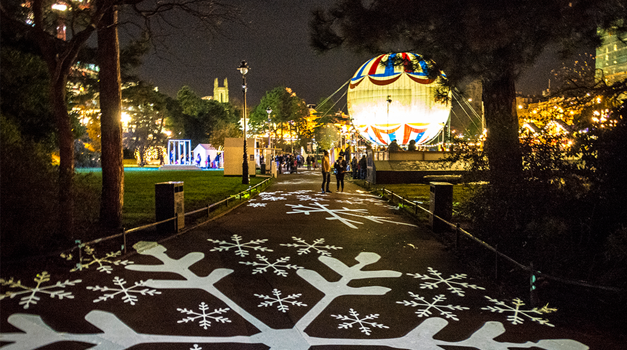 A festive nighttime scene with a pathway illuminated by snowflake patterns and a large, decorated sphere in the background.