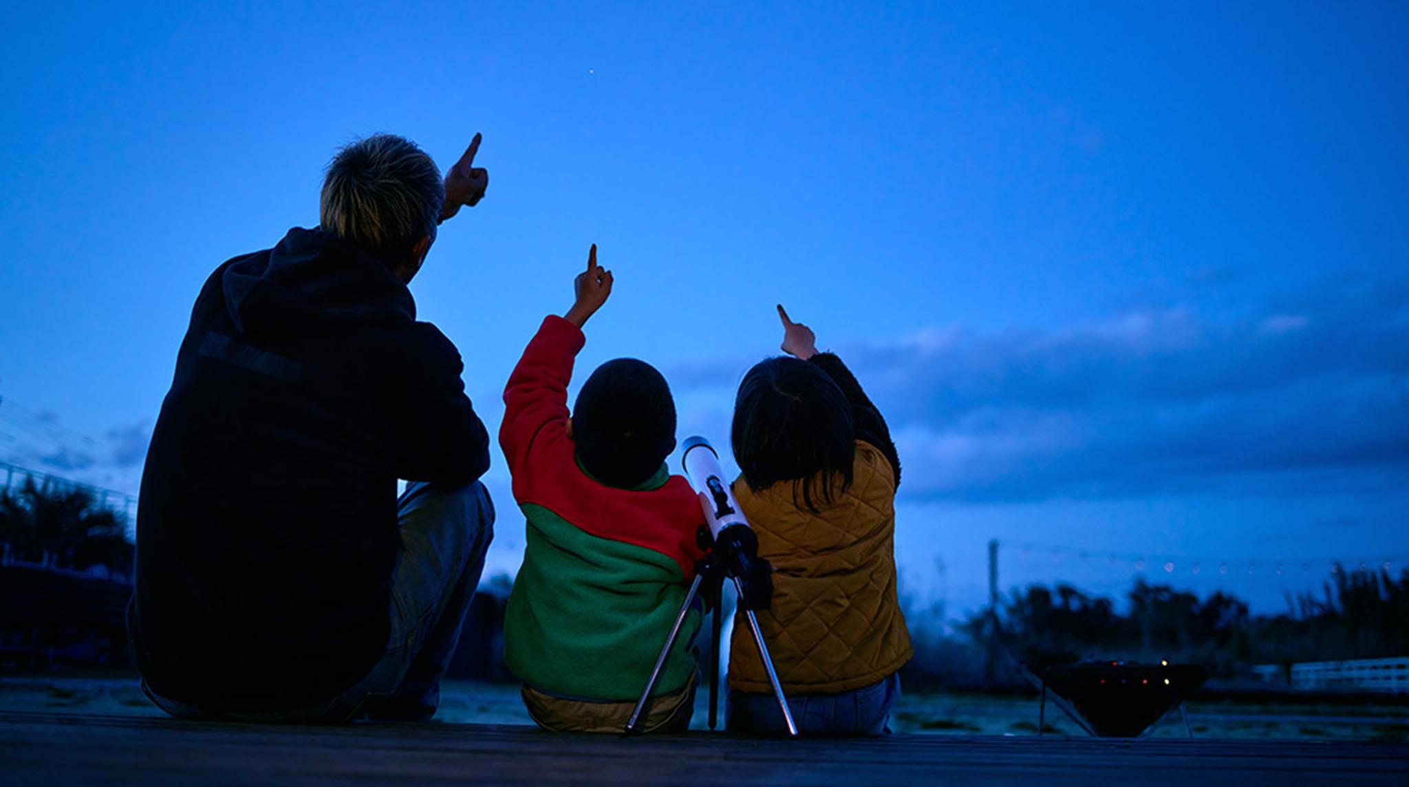 Three people sitting on a wooden deck at dusk, pointing at the sky with a telescope nearby