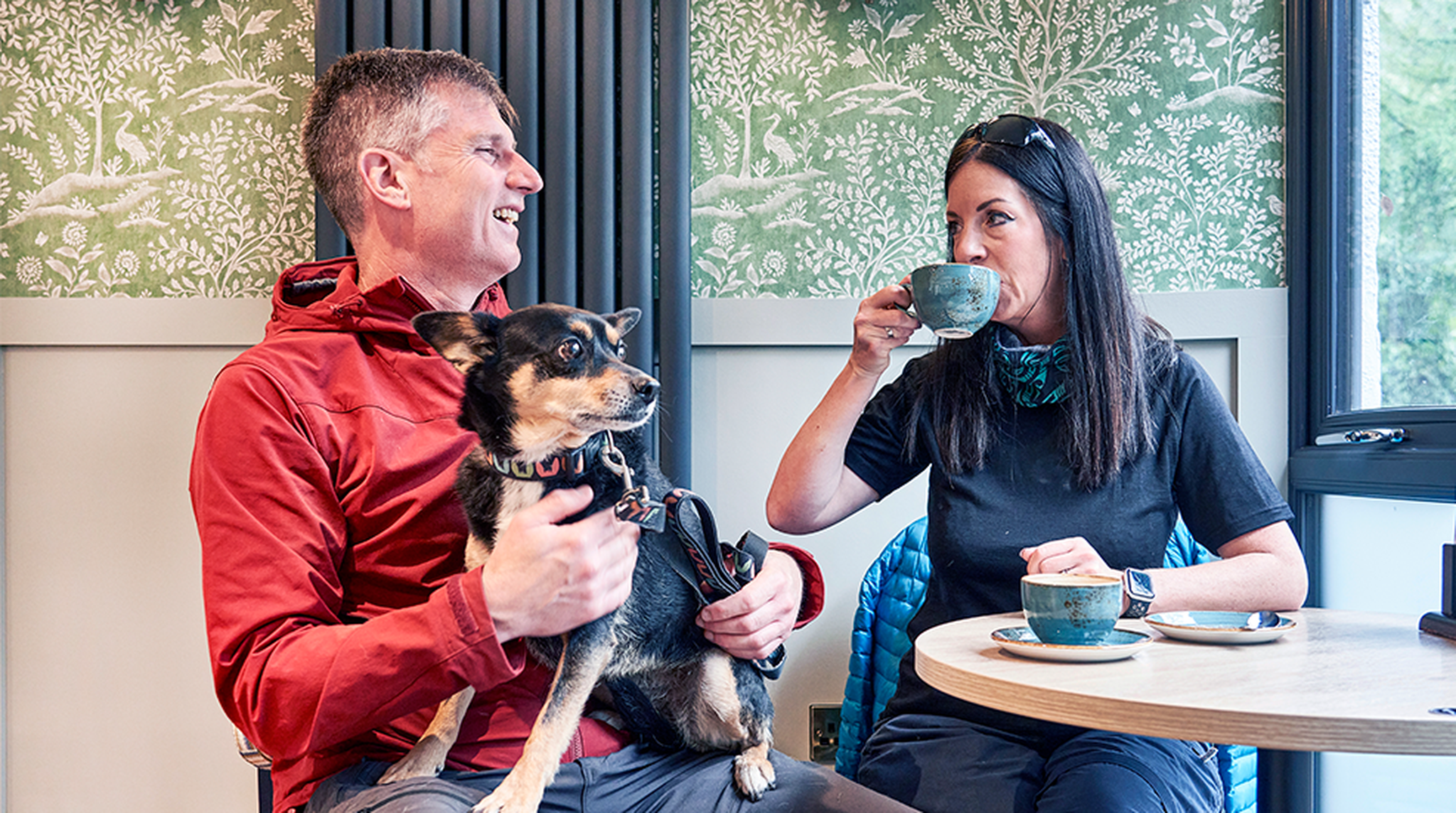A man holding a dog and a woman sipping tea share a moment at a café table, with green decorative wallpaper in the background.