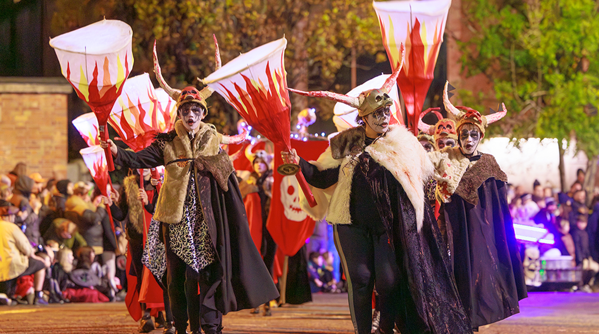 A nighttime street scene shows a procession of performers dressed in horned headpieces, face paint, fur-trimmed capes, and dark costumes in Derry