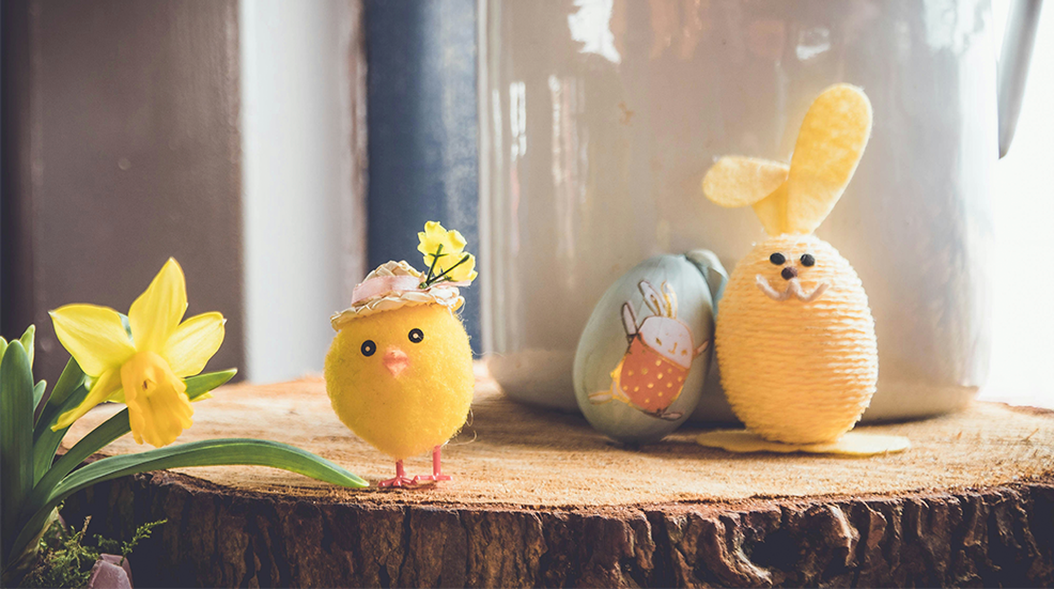 Cute Easter decorations include a plush chick with a hat, daffodil, bunny figure, and decorated egg on a wooden surface.