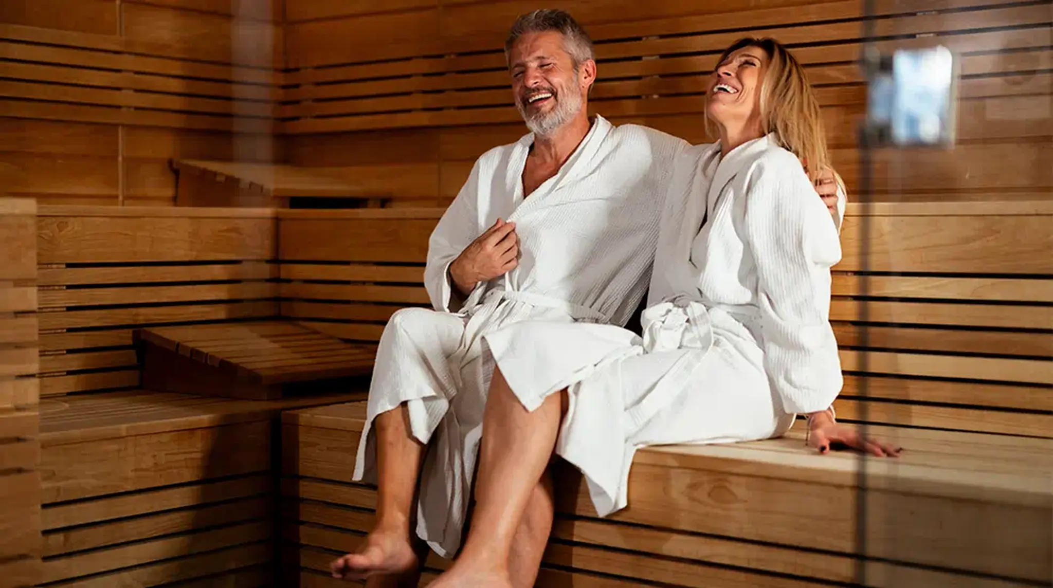 A smiling couple in white robes sit on a wooden bench in a sauna
