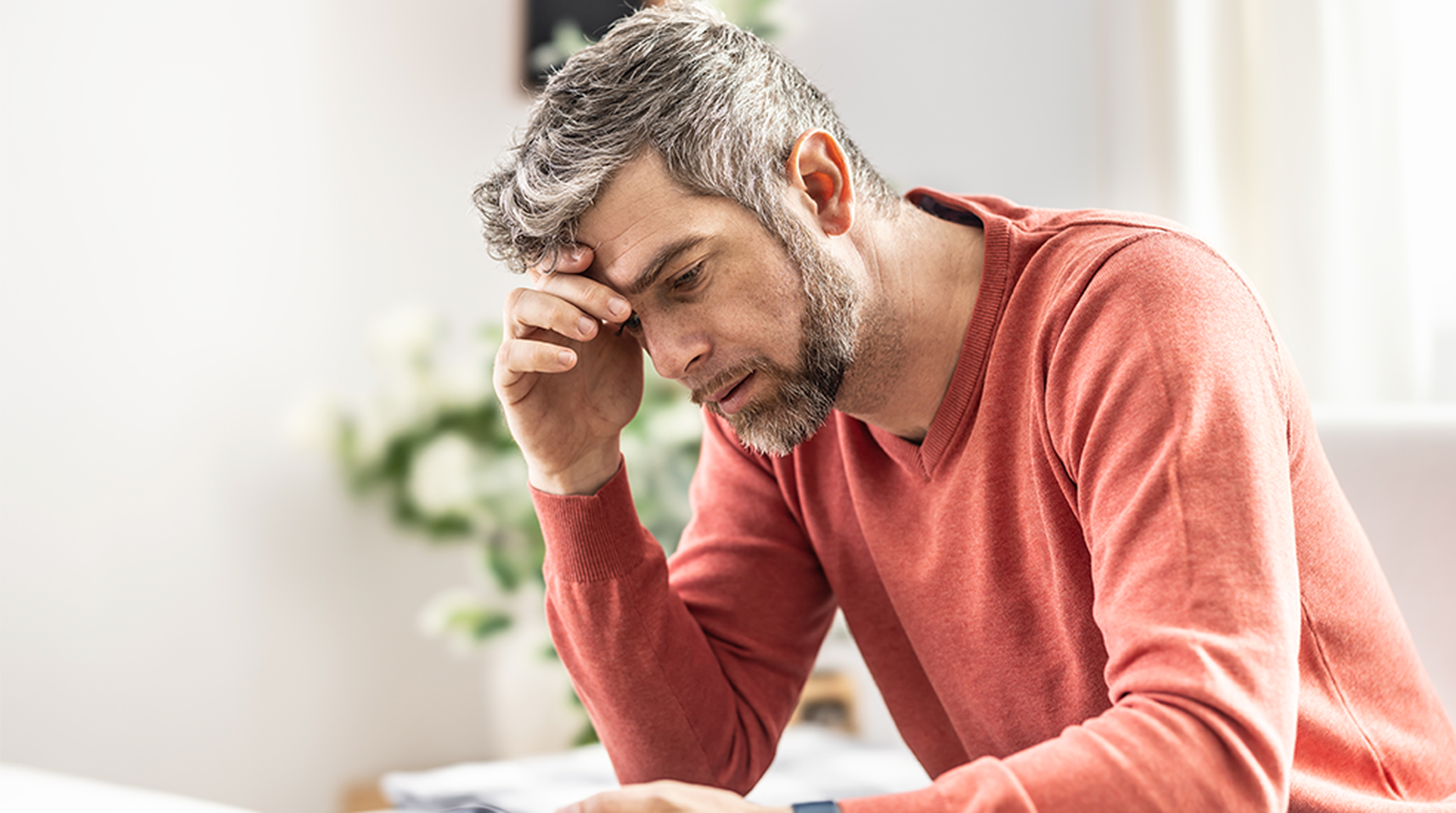 Man in a red sweater looks concerned while sitting indoors, with one hand on his forehead