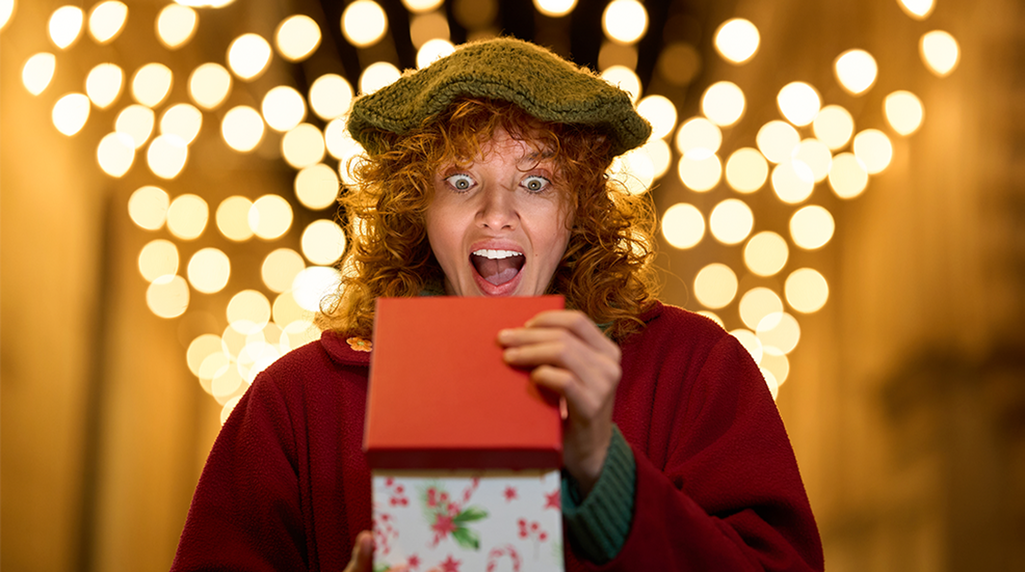 Person with curly hair excitedly opening gift box under bright festive lights.