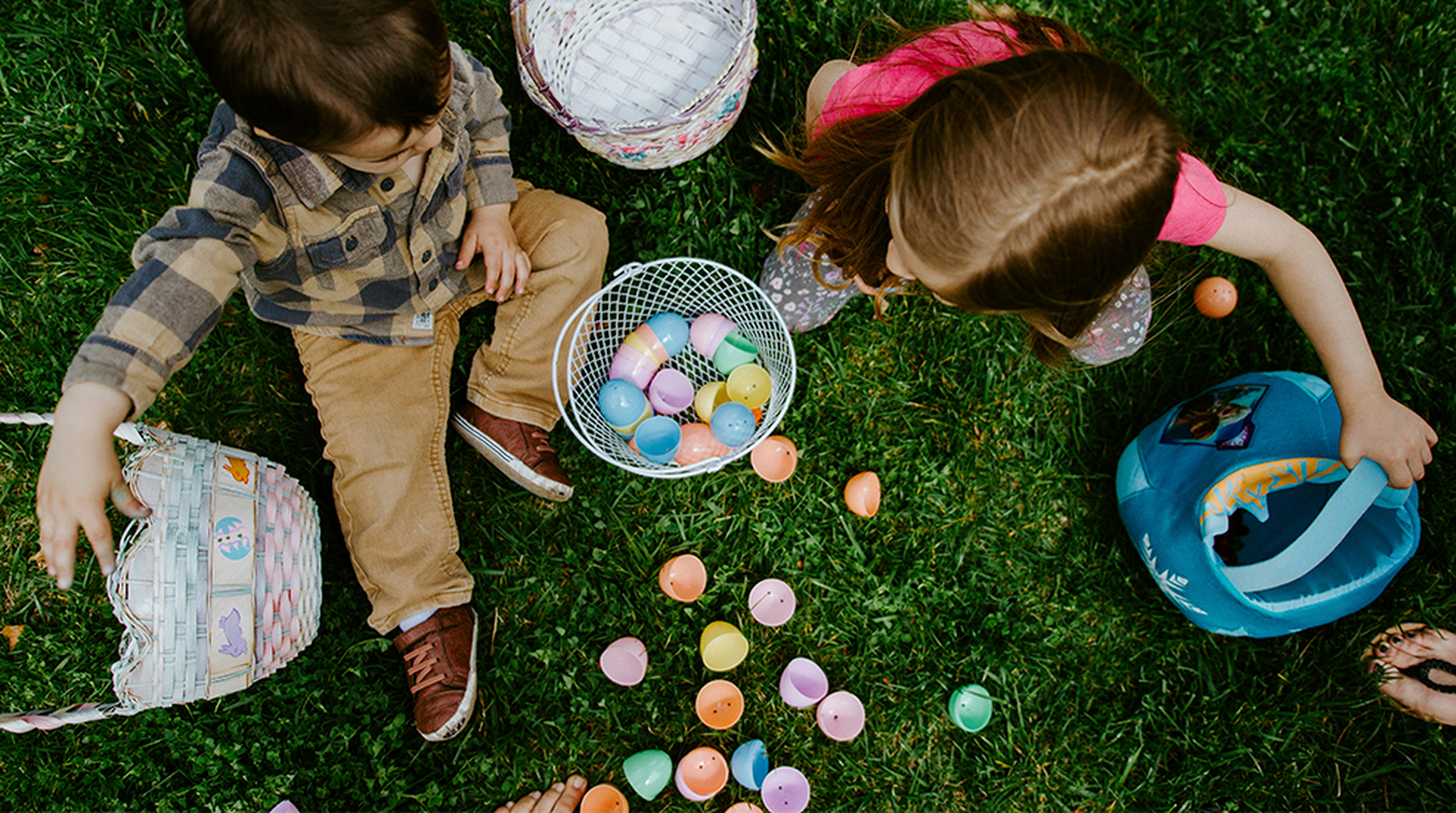 Two children sitting on grass with baskets, gathering colorful plastic eggs, possibly during an Easter egg hunt.