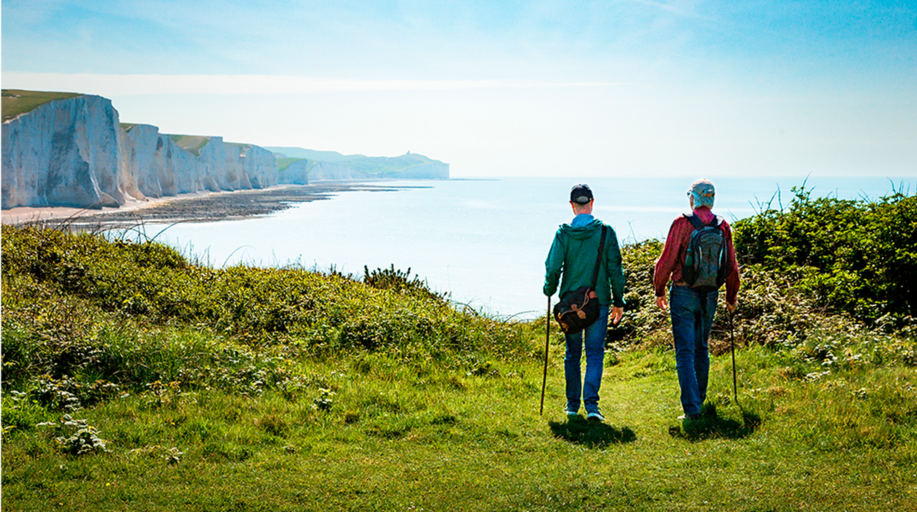 Two hikers walking on a grassy path towards white cliffs by the sea under a clear blue sky.