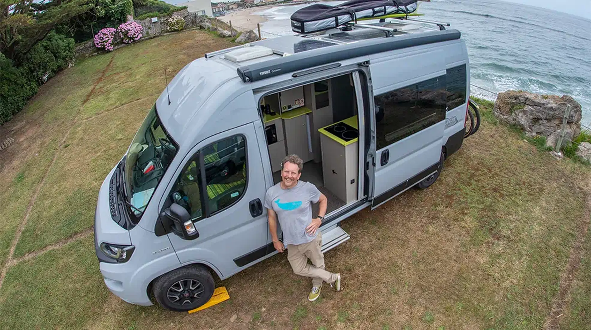 Man leaning on a grey camper van parked on grass near a beach