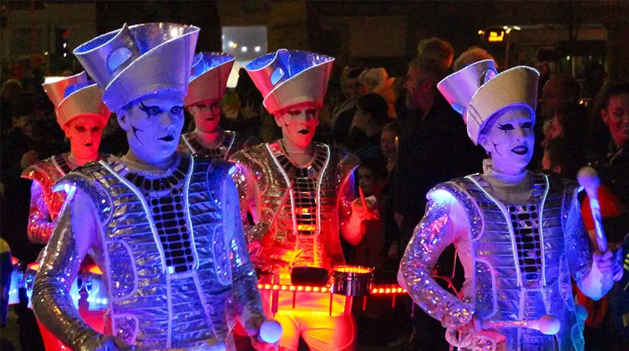 Performers in futuristic costumes with glowing lights play instruments in a night time street parade