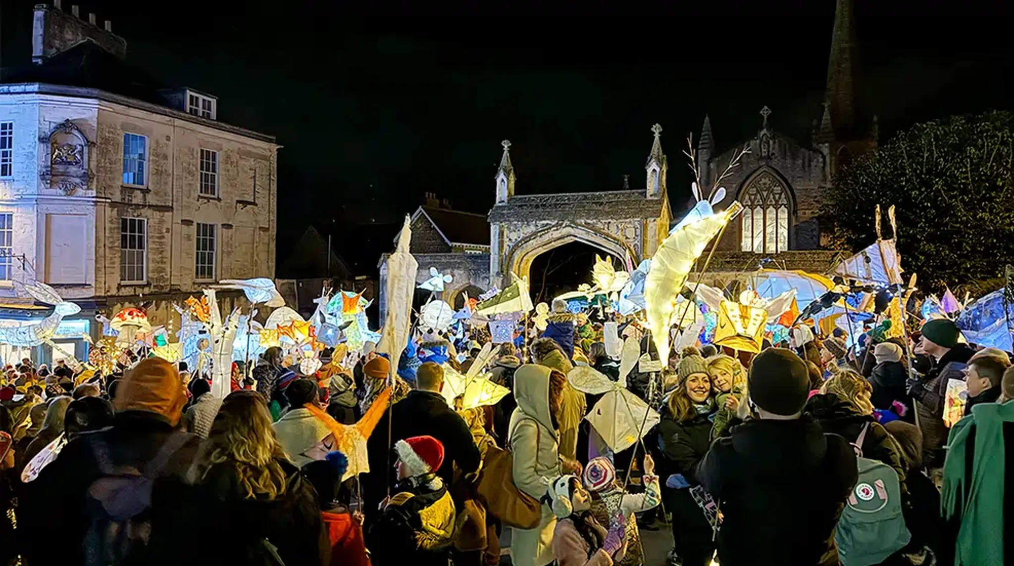 A festive outdoor night time crowd holds glowing lanterns and animal-shaped sculptures