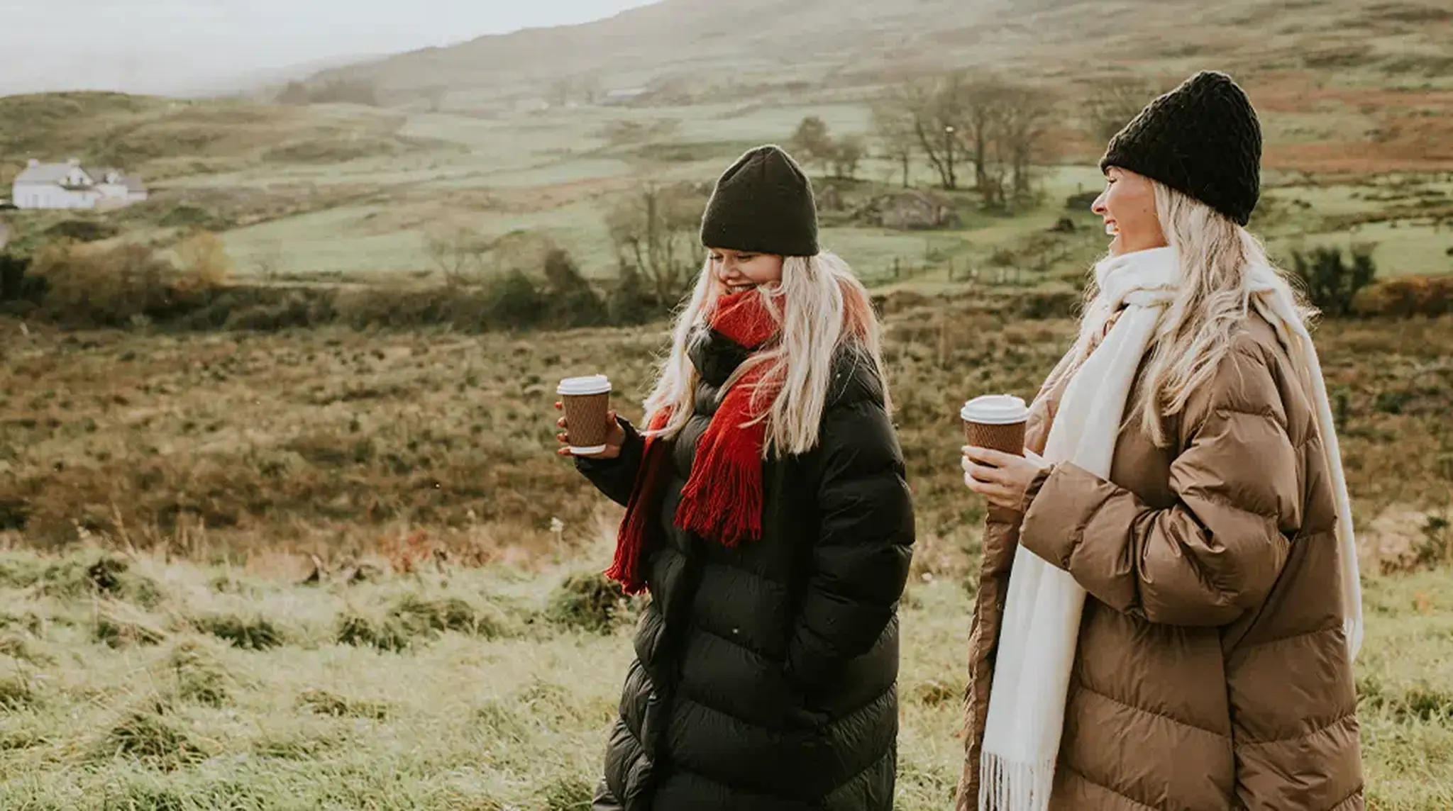 Two women in winter coats and beanies, holding cups, stand in a grassy landscape