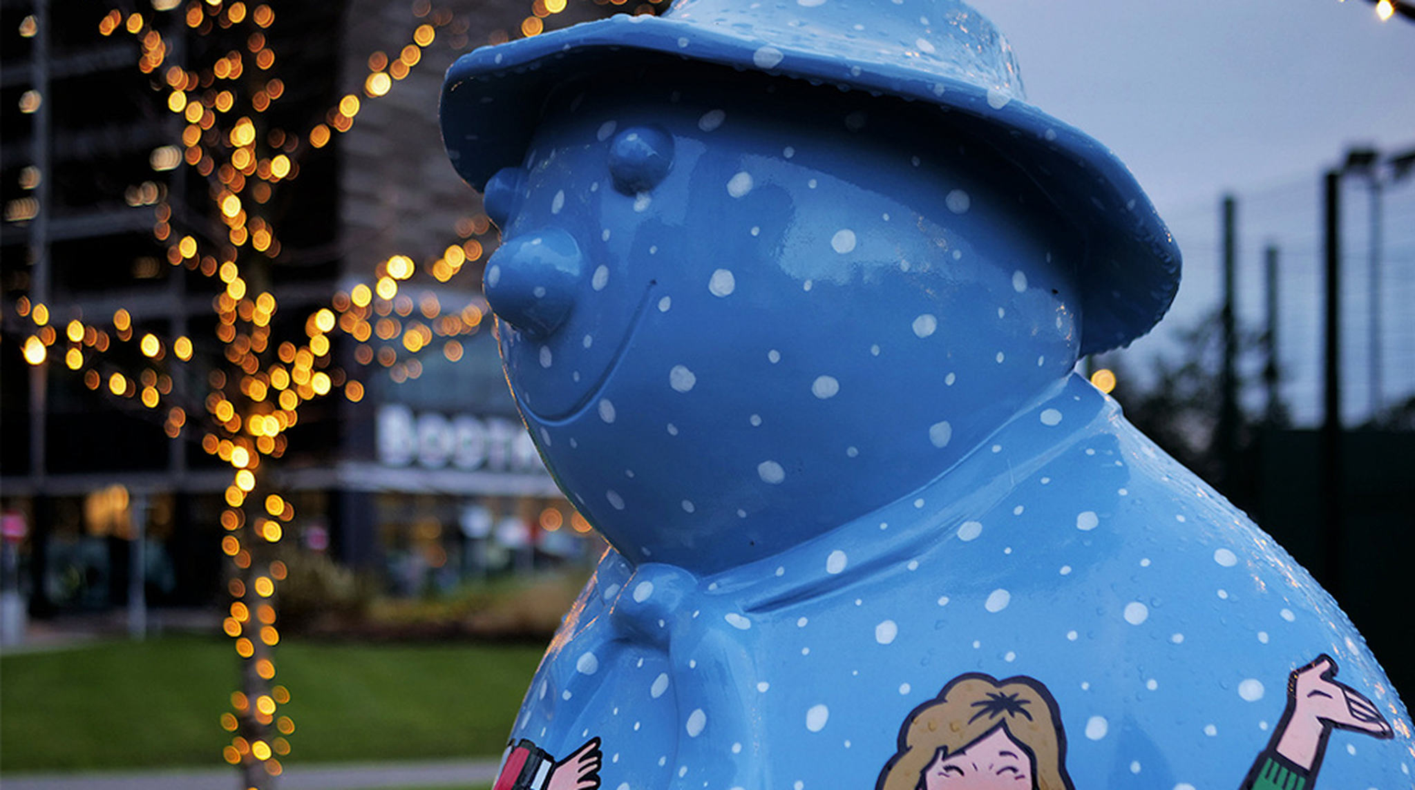 A large blue snowman statue with a hat is adorned with white polka dots, set against a backdrop of festive string lights.