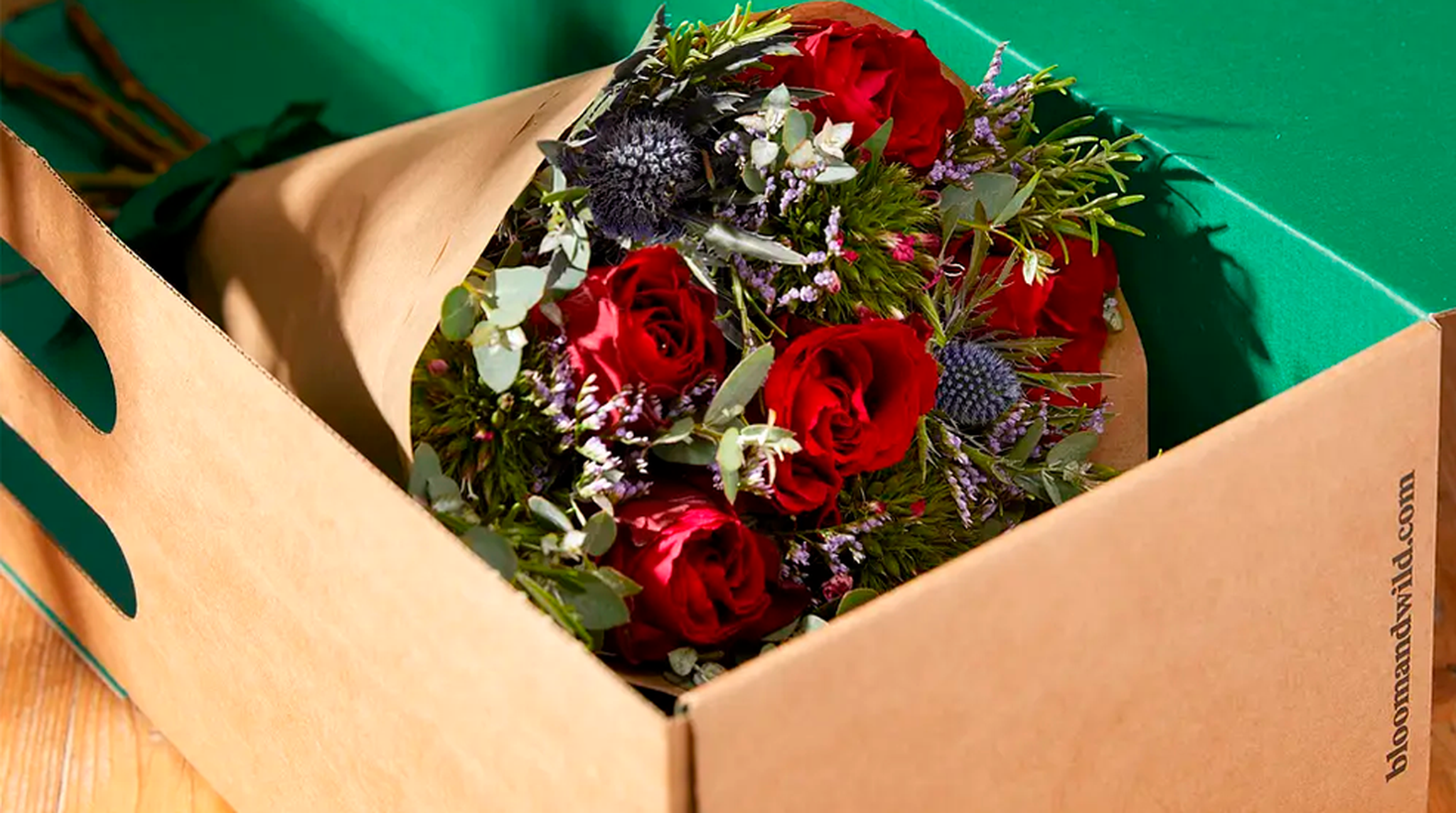 Bouquet of red roses with greenery and purple flowers inside an open cardboard box.