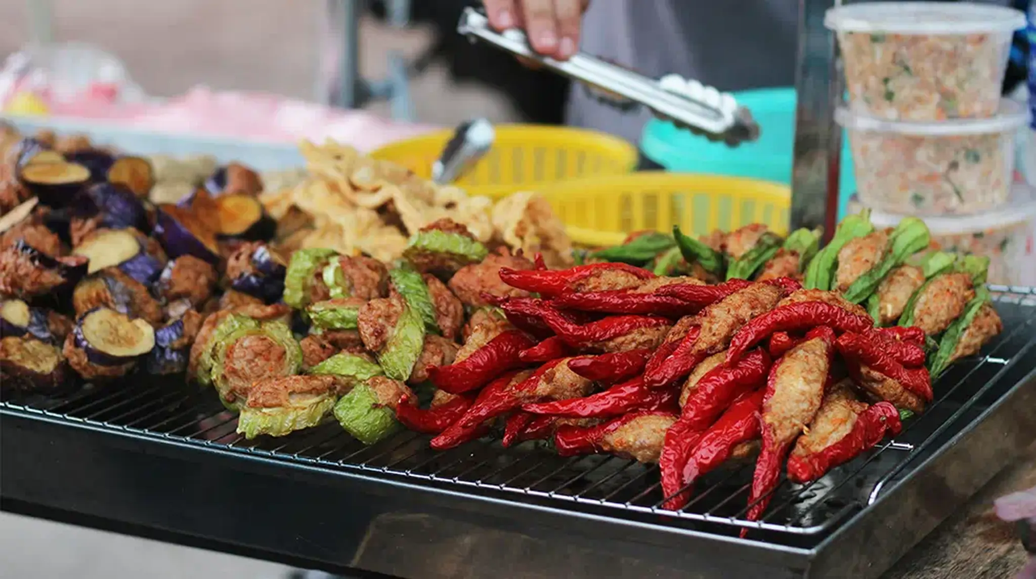 A street food stall with fried vegetables and stuffed red and green peppers arranged on a grill