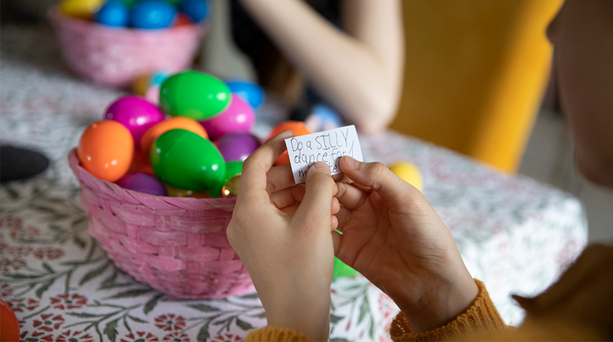 Hands holding a note saying, "Do a SILLY dance," with colorful plastic eggs in a pink basket on a patterned tablecloth in the background.