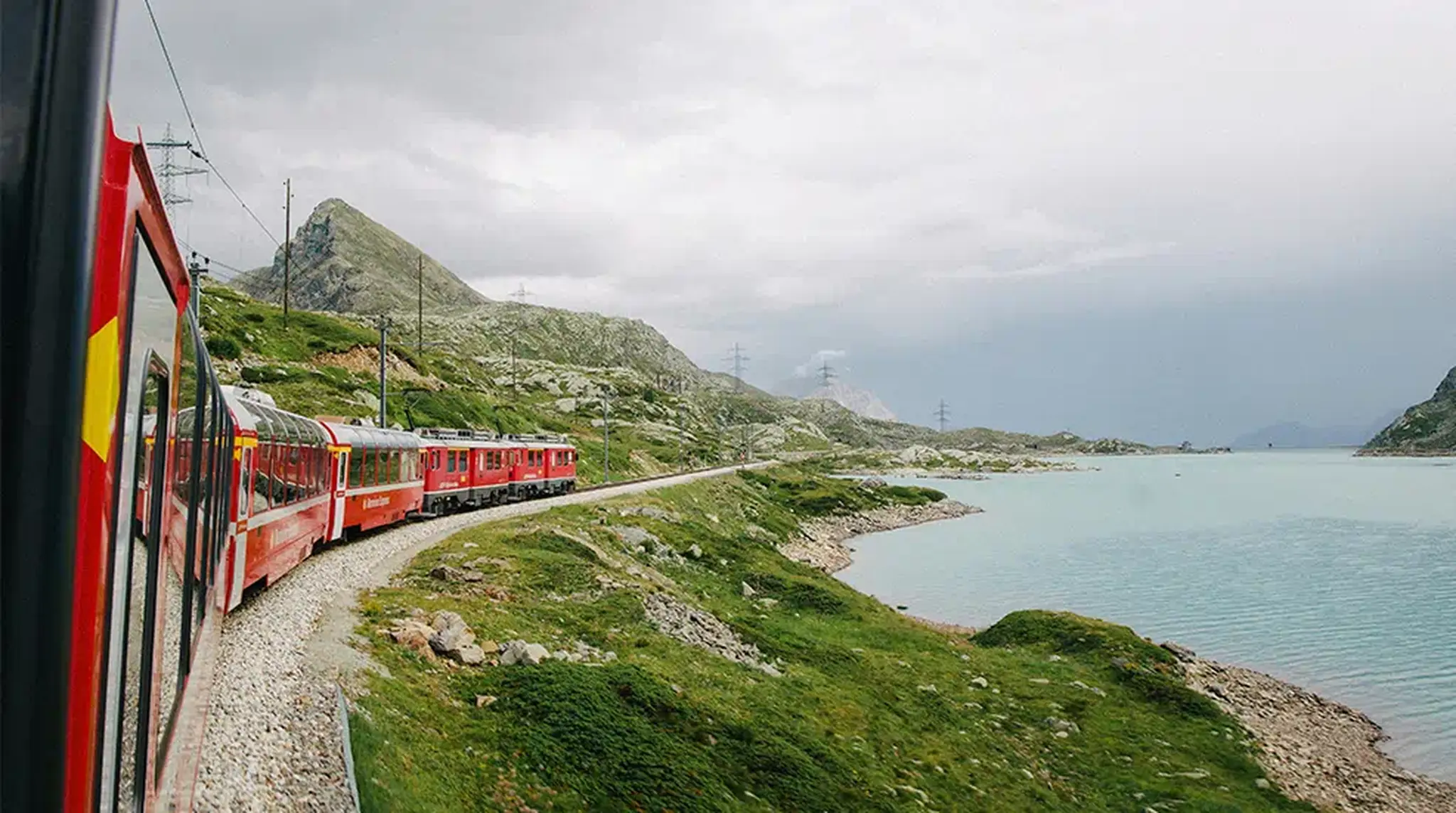 A red train travels along a curved track beside a serene lake and green hills