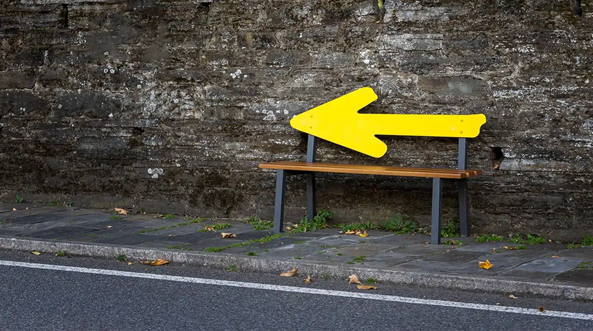A bench with a large, yellow arrow-shaped backrest against a stone wall beside a road.