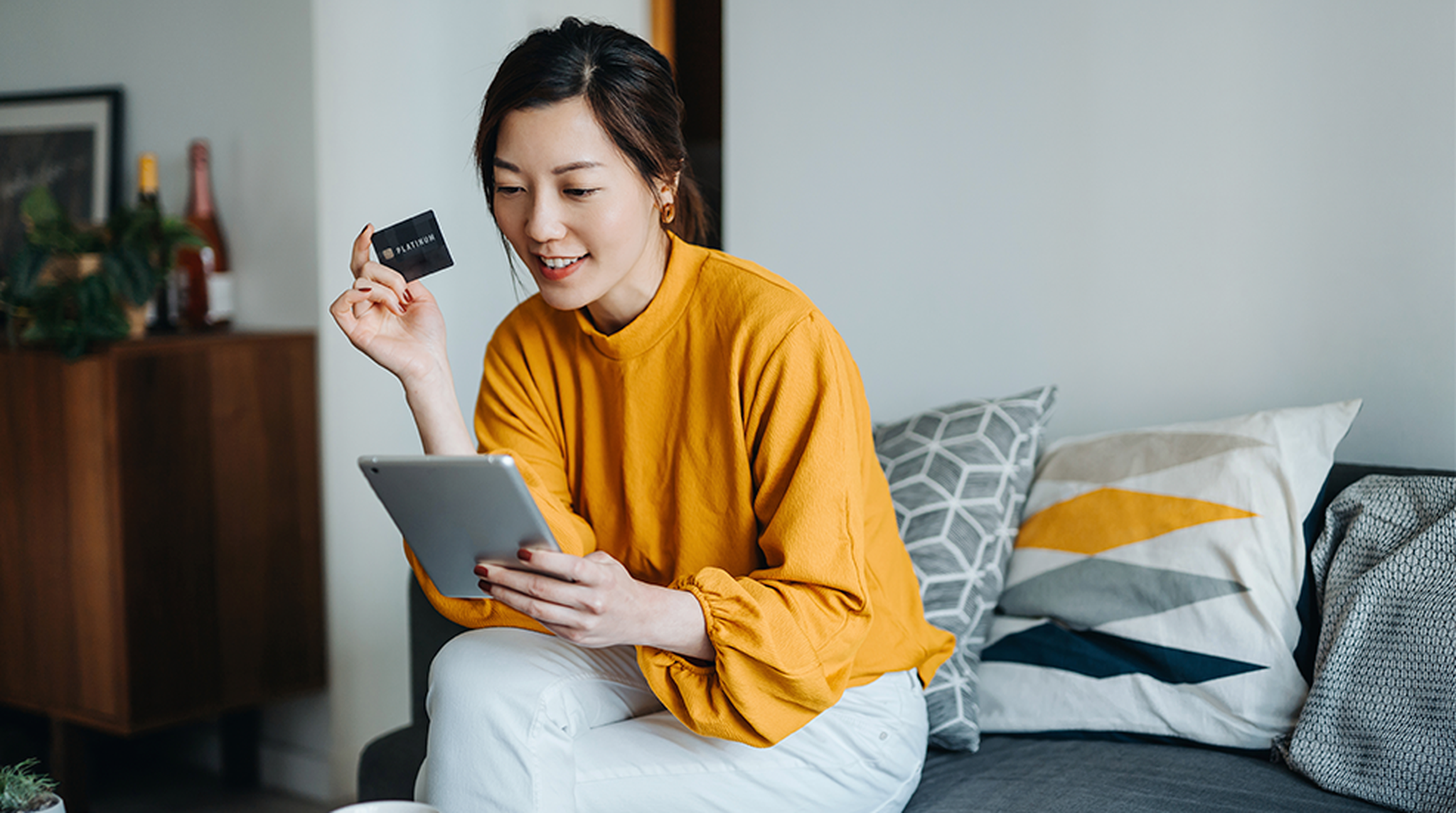Woman in a yellow blouse holds a credit card and uses a tablet while sitting on a sofa