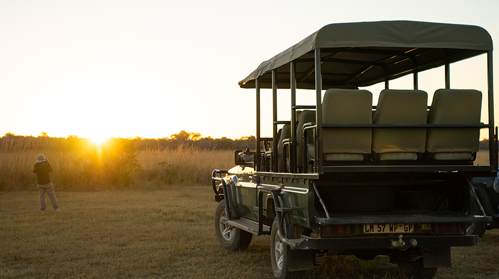 Safari jeep parked on grassland at sunset, with a person taking a photo of the scene.