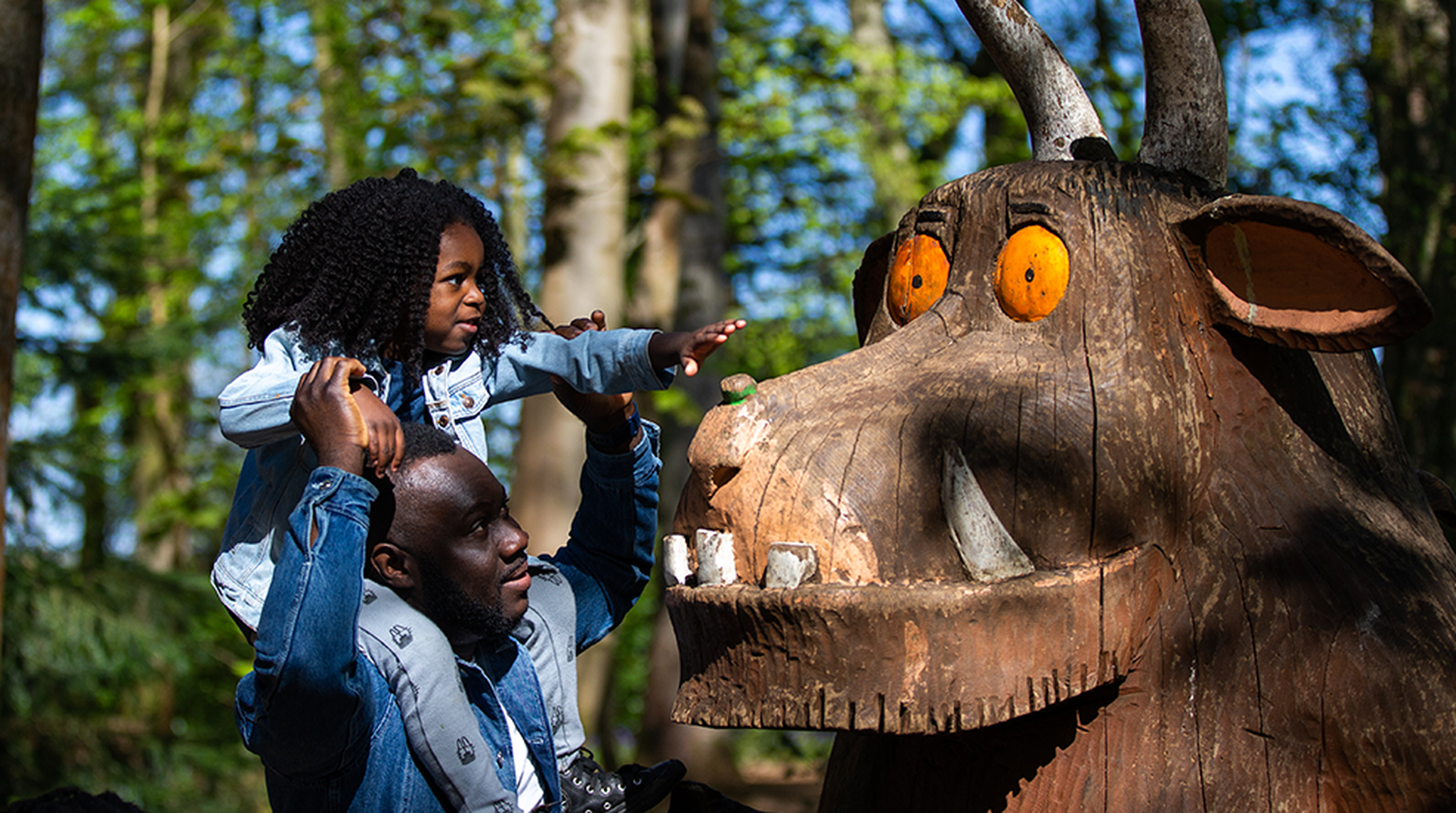 A black father with his daughter on his shoulders, reaching out to the Gruffalo statue in Westonbirt Aboretum