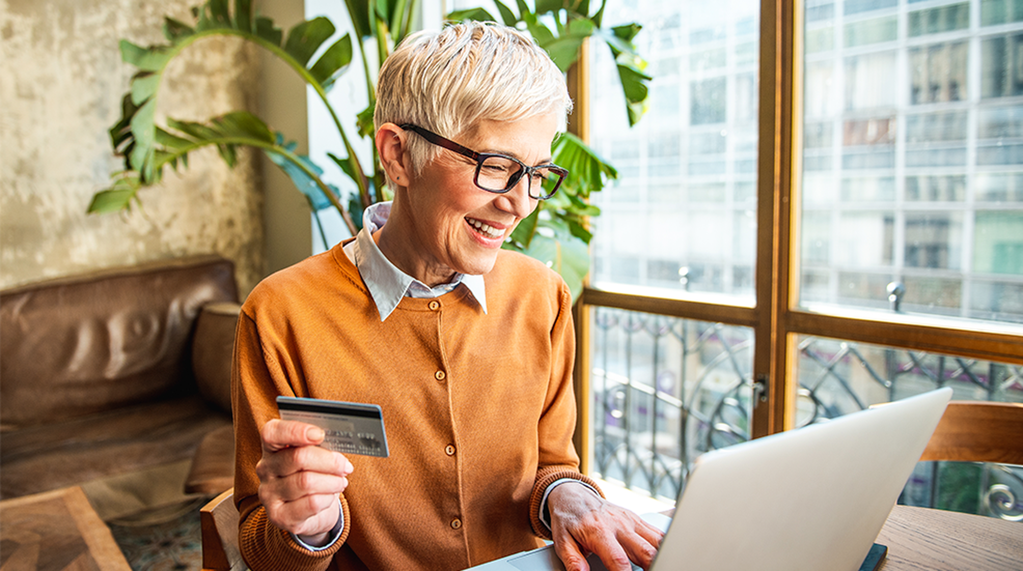 Older woman with short hair smiling, holding a credit card, and using a laptop