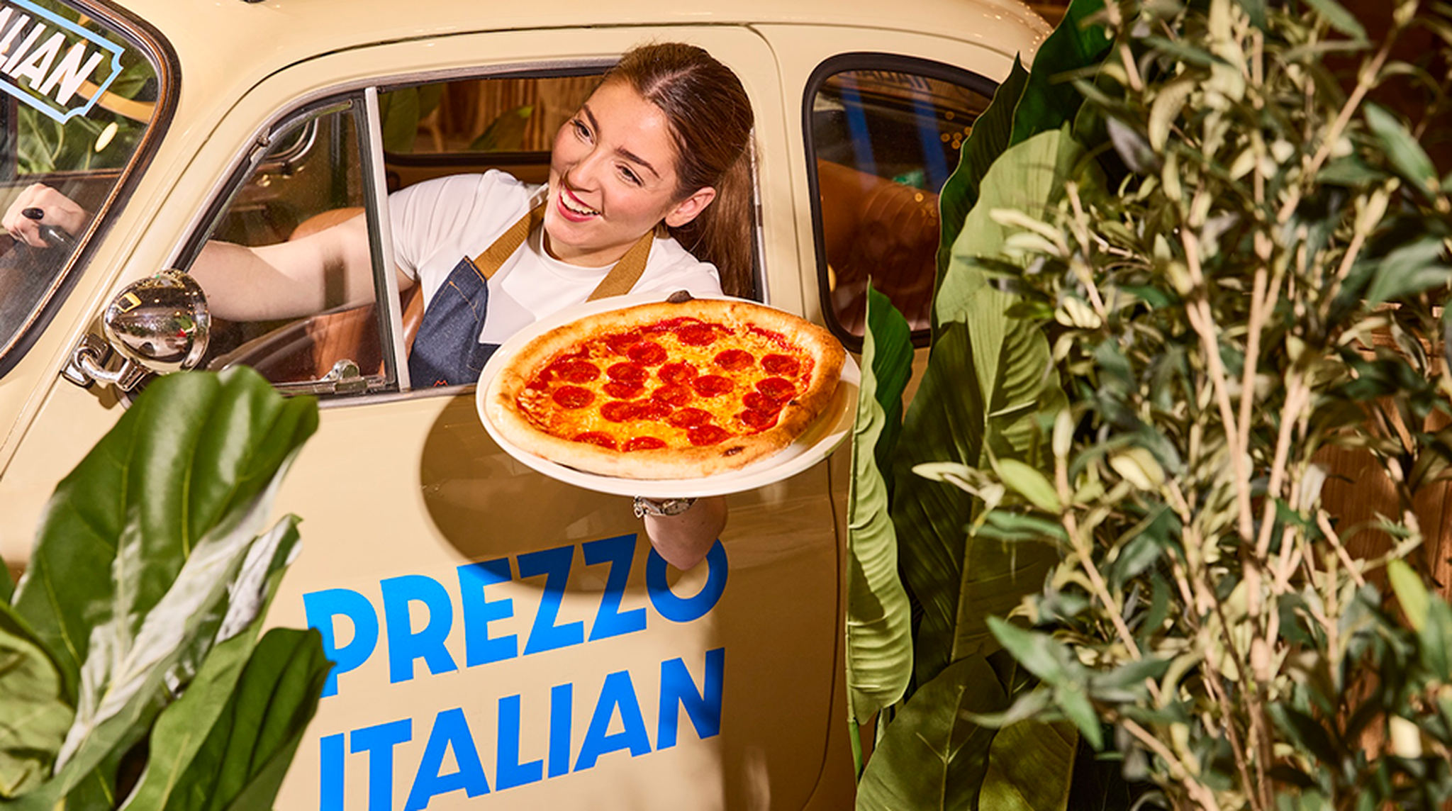 Woman hanging outside of classic car window branded with Prezzo Italian holding a pizza