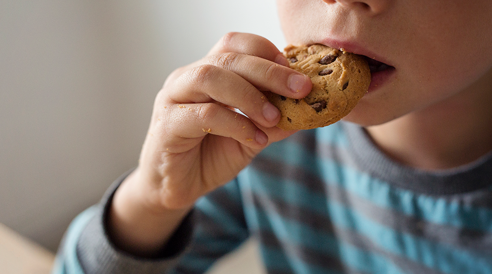 A child in a striped shirt takes a bite of a chocolate chip cookie, with focus on the cookie and hand.