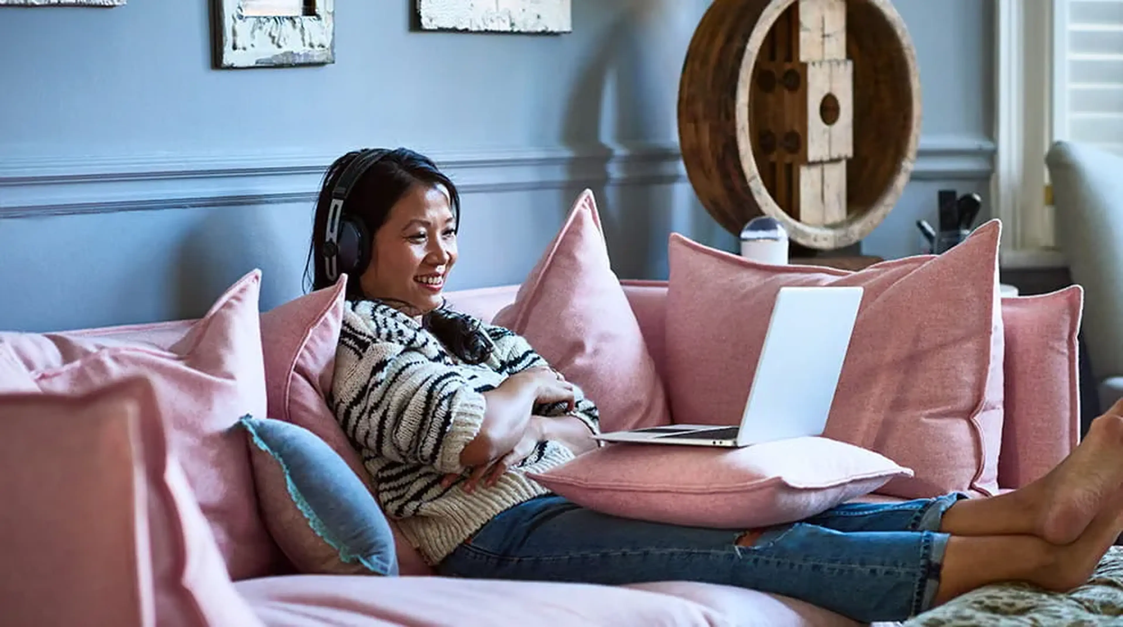 Woman sitting on a pink sofa, wearing headphones and smiling at a laptop