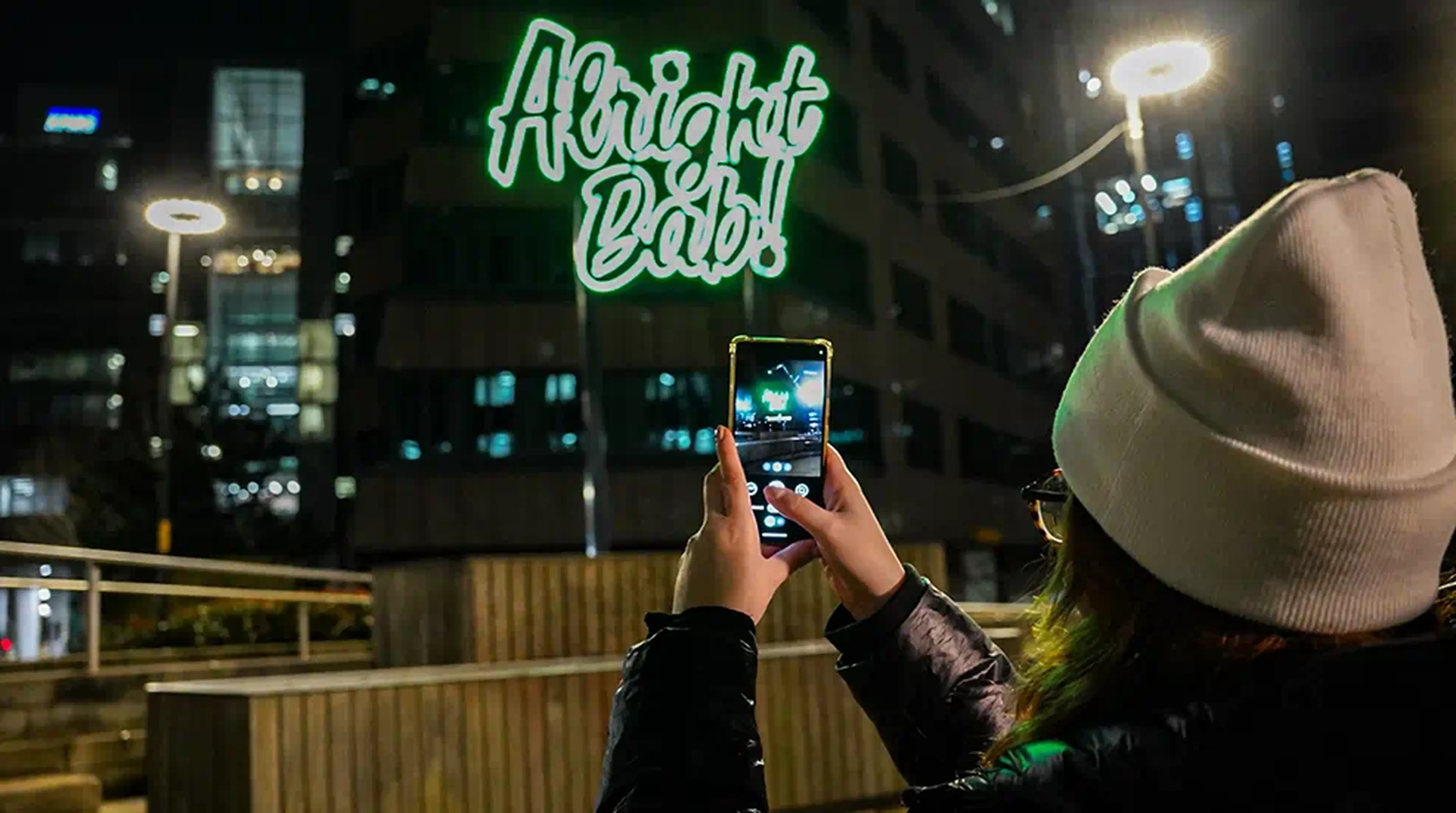 Person taking a photo of a neon sign reading "Alright Bab!" on a building