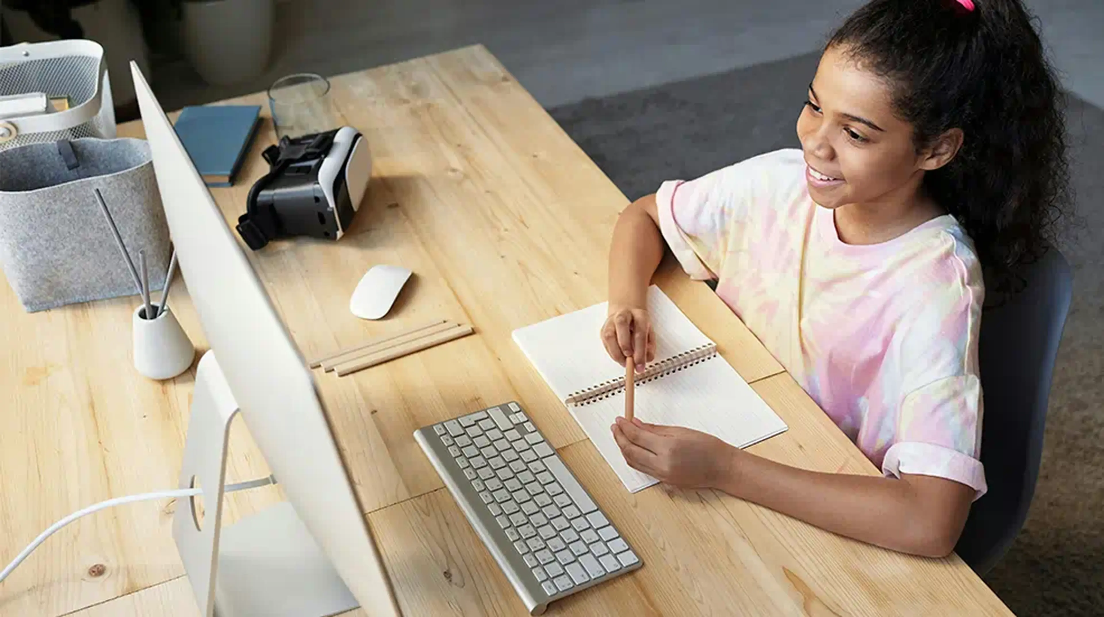 Smiling girl in a tie-dye shirt writing in a notebook at a desk with a computer