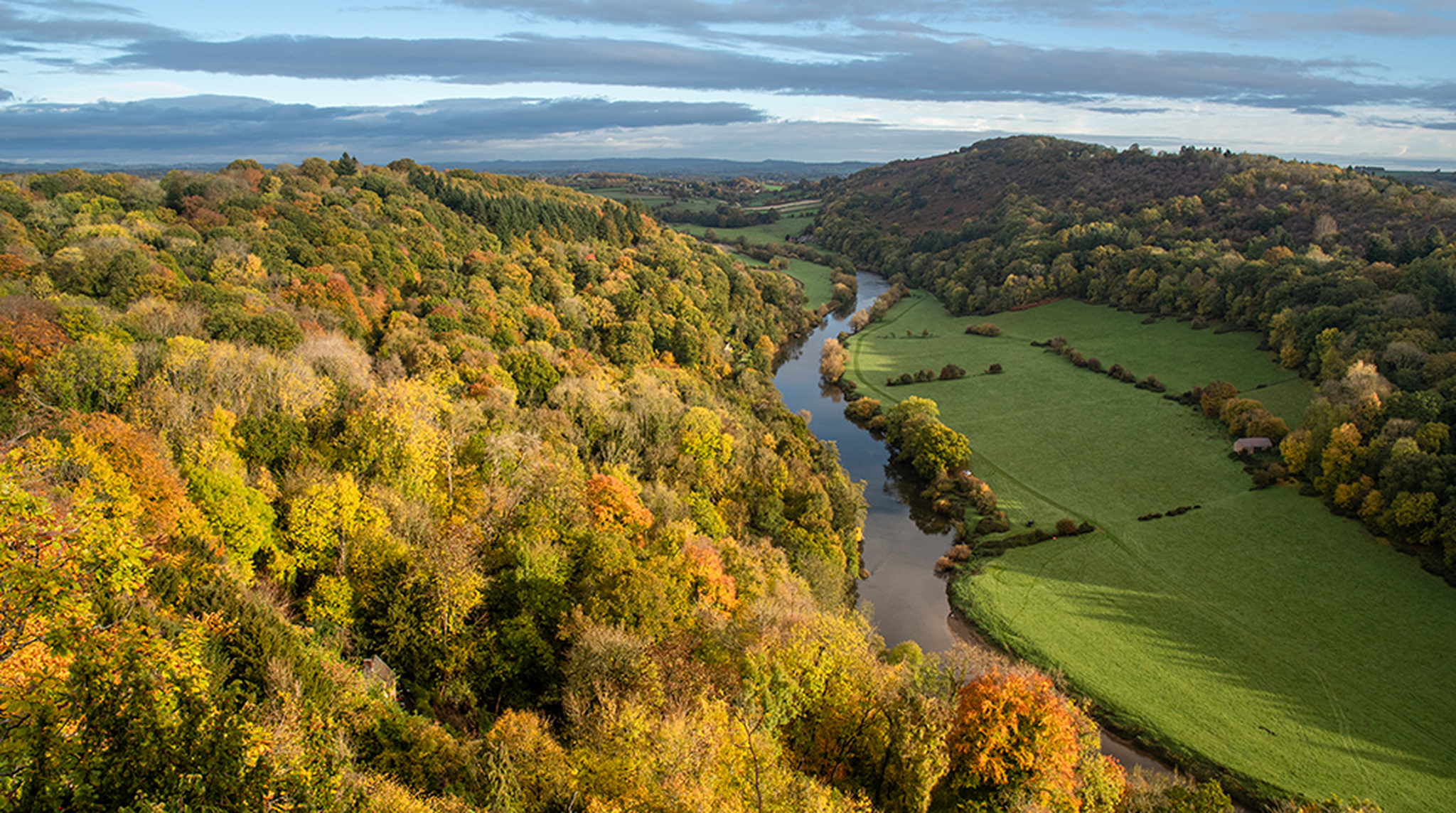High angle view of a winding river flowing through a lush green and autumnal forest valley under a cloudy sky, Forest of Dean