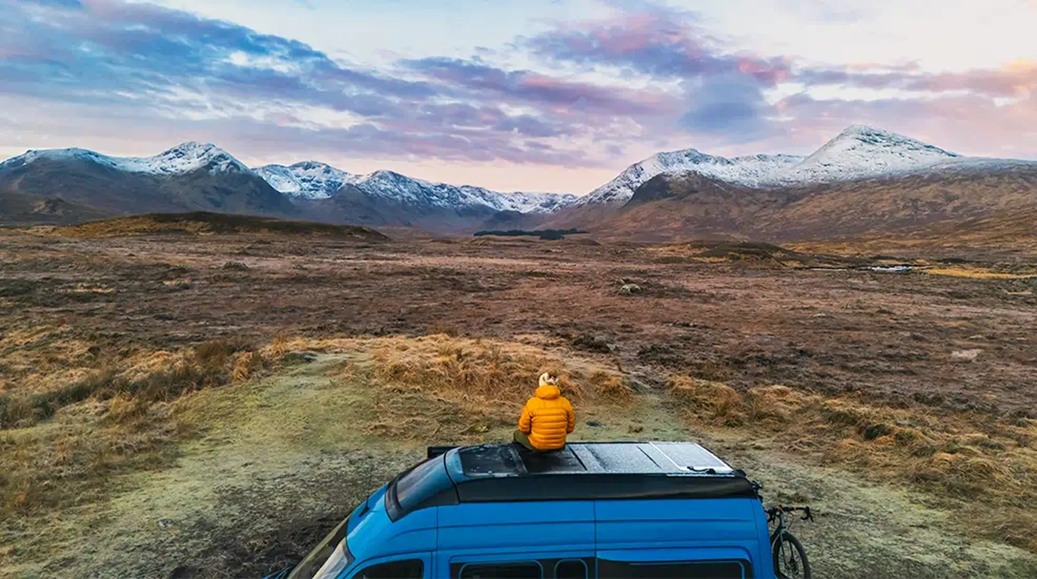 Person in a yellow jacket sits on a blue van roof, overlooking a vast landscape