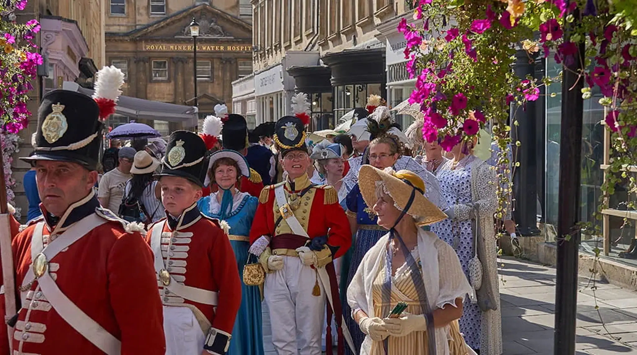 People in historical costumes stroll through a street with hanging flowers
