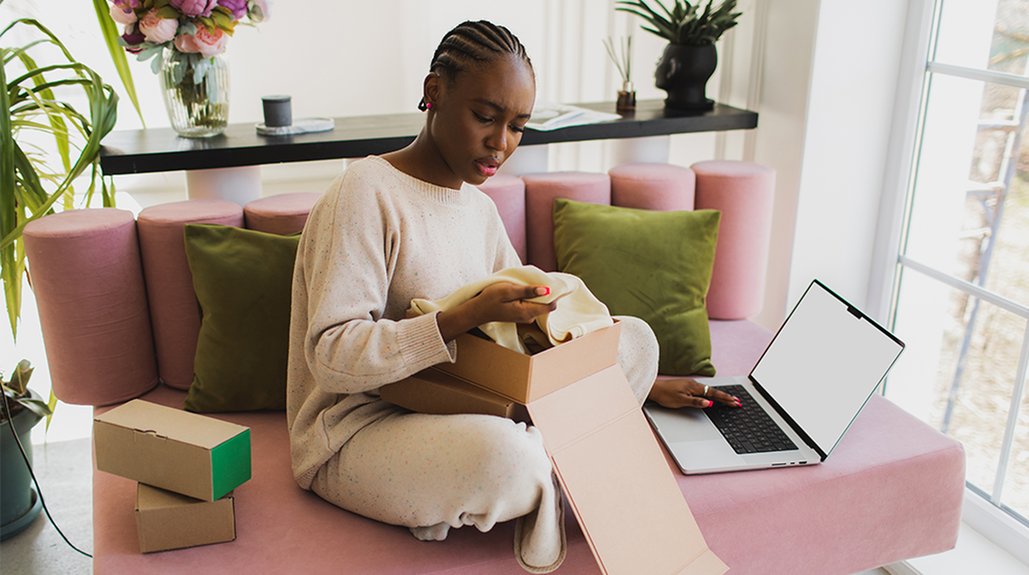 A person sitting on a pink sofa at home, looking at a garment from an open box while using a laptop, surrounded by green cushions and plants