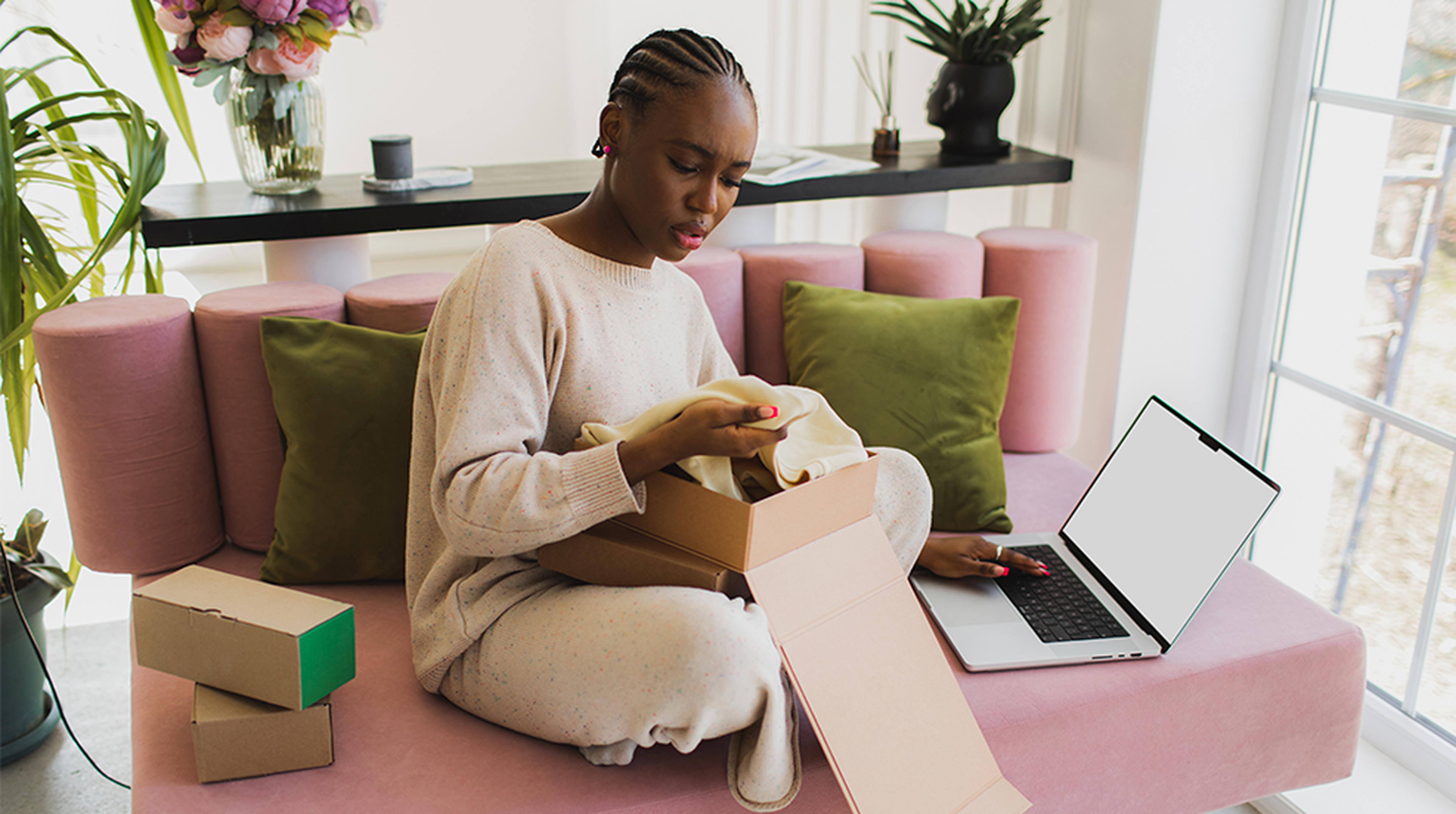 A person sitting on a pink sofa at home, looking at a garment from an open box while using a laptop, surrounded by green cushions and plants