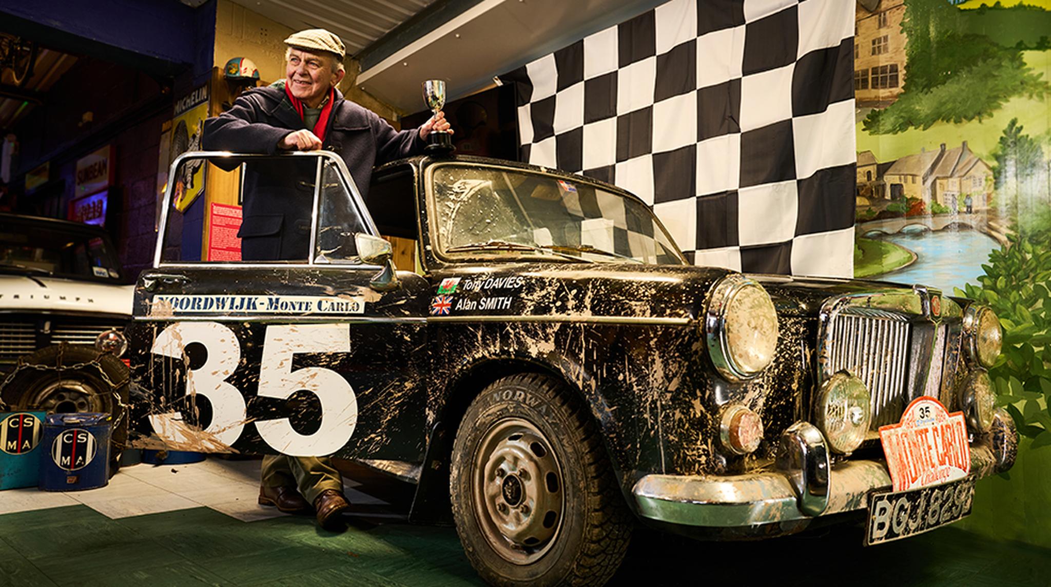 Elderly man with a trophy stands beside a classic, mud-splattered Monte Carlo rally car adorned with racing number 35 and checkered flag.
