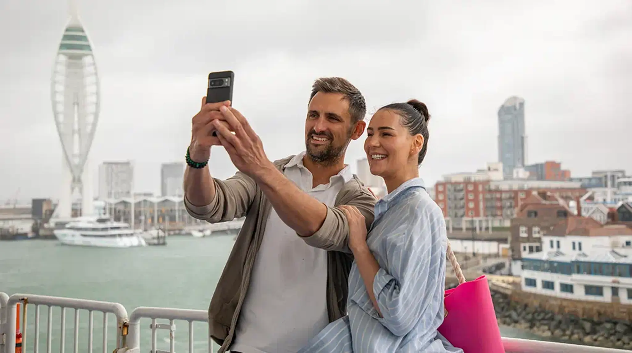 A couple takes a selfie on a waterfront, with a cityscape and tall spire in the background on a cloudy day.