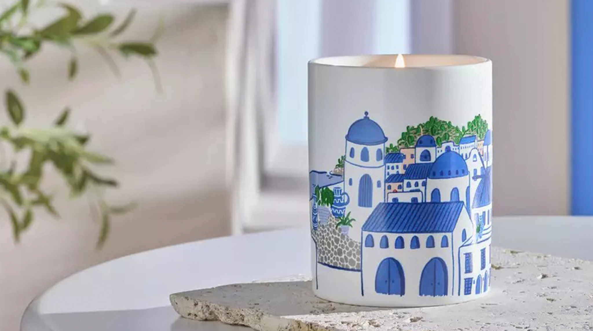 Lit candle in a decorative holder featuring a blue and white illustration of traditional Mediterranean architecture