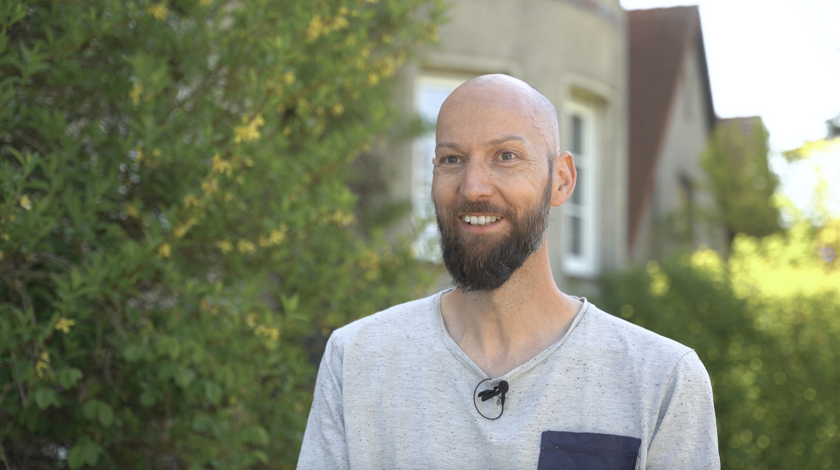 Smiling man with a beard, wearing a gray shirt with a microphone, stands outdoors in front of greenery and a house.