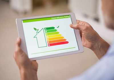 Person hält ein Tablet, auf dem eine Tabelle zur Bewertung der Energieeffizienz von Häusern mit Kategorien von A (grün) bis G (rot) angezeigt wird.