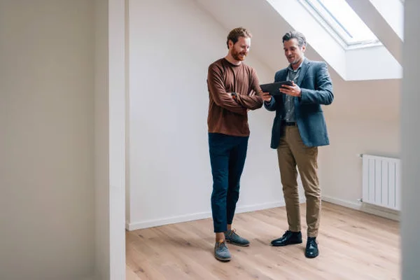 Zwei Männer stehen in einem hellen Raum mit Holzboden und diskutieren etwas auf einem Tablet. Der eine trägt einen Pullover, der andere einen Blazer.