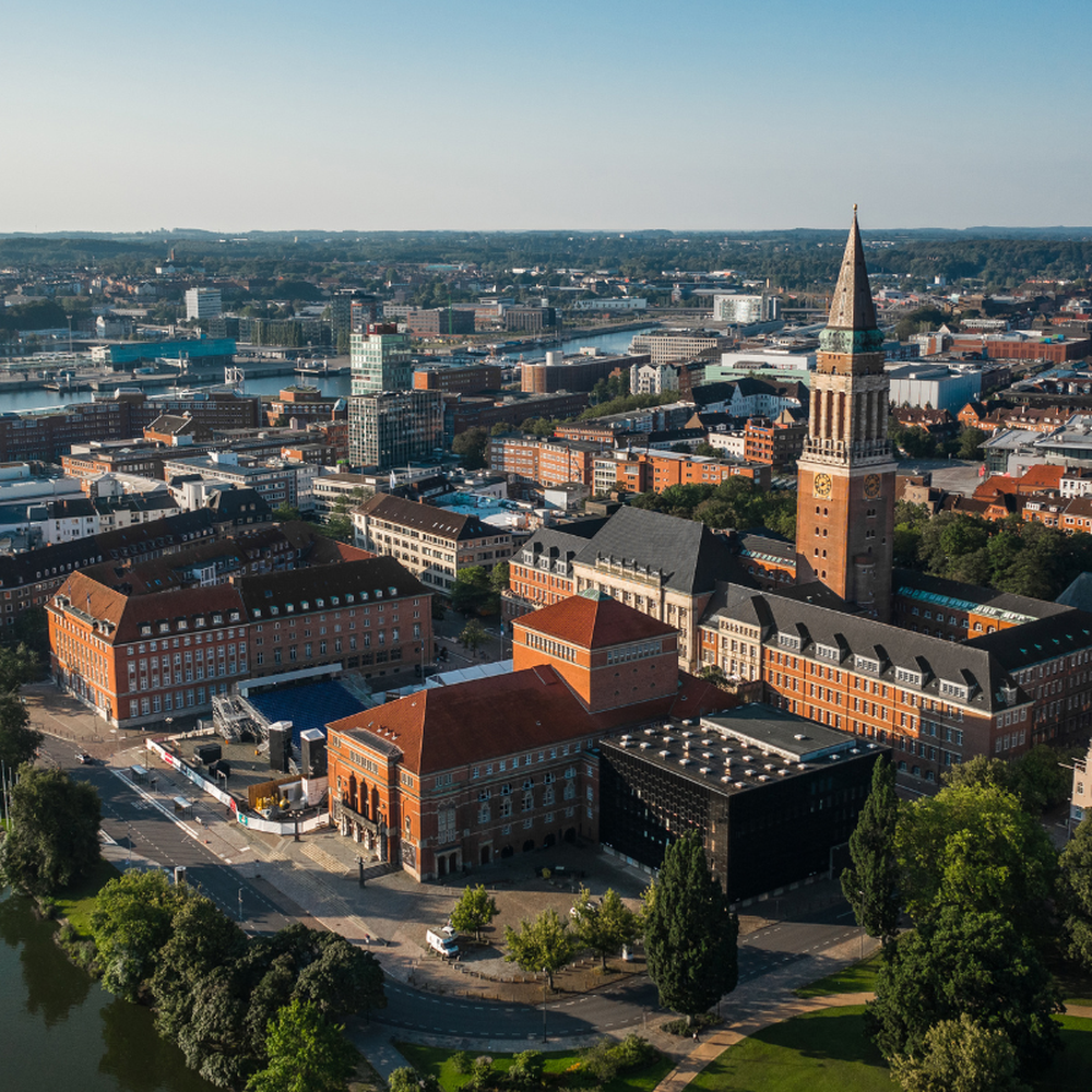 Luftaufnahme des Kieler Rathauses mit Turm und Rathausplatz, im Hintergrund die Förde und Teile der Innenstadt bei klarem Wetter.