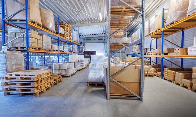 Prostorný interiér skladu se stohovanými paletami, krabicemi a policemi plnými zboží. Jasné osvětlení a uspořádaný layout.