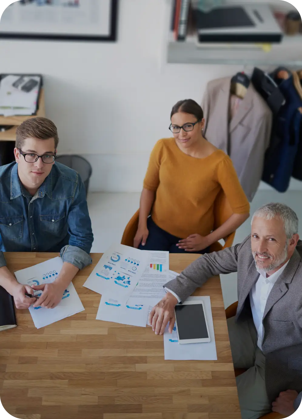 Tři lidé sedí u stolu v kanceláři, na stole jsou papíry s grafy, tablet a blok